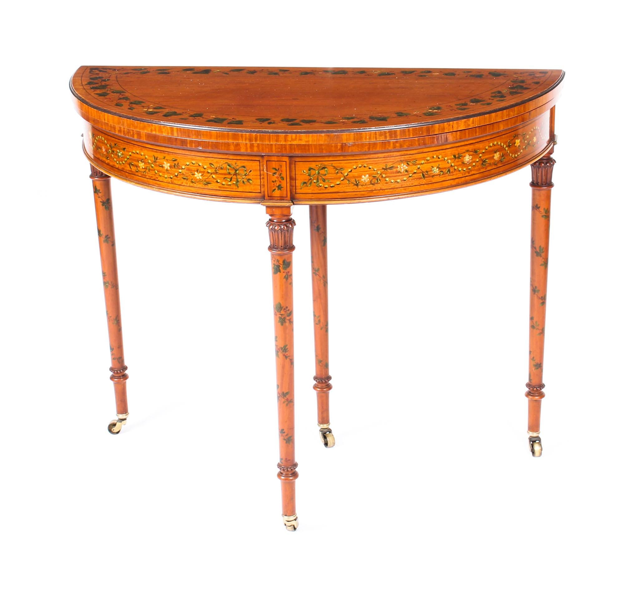 This is a wonderful antique hand painted satinwood Sheraton Revival demilune card table, circa 1890 in date.

The folding top enclosing an inset baize lined playing surface. It is decorated throughout with hand painted bands of trailing vines and