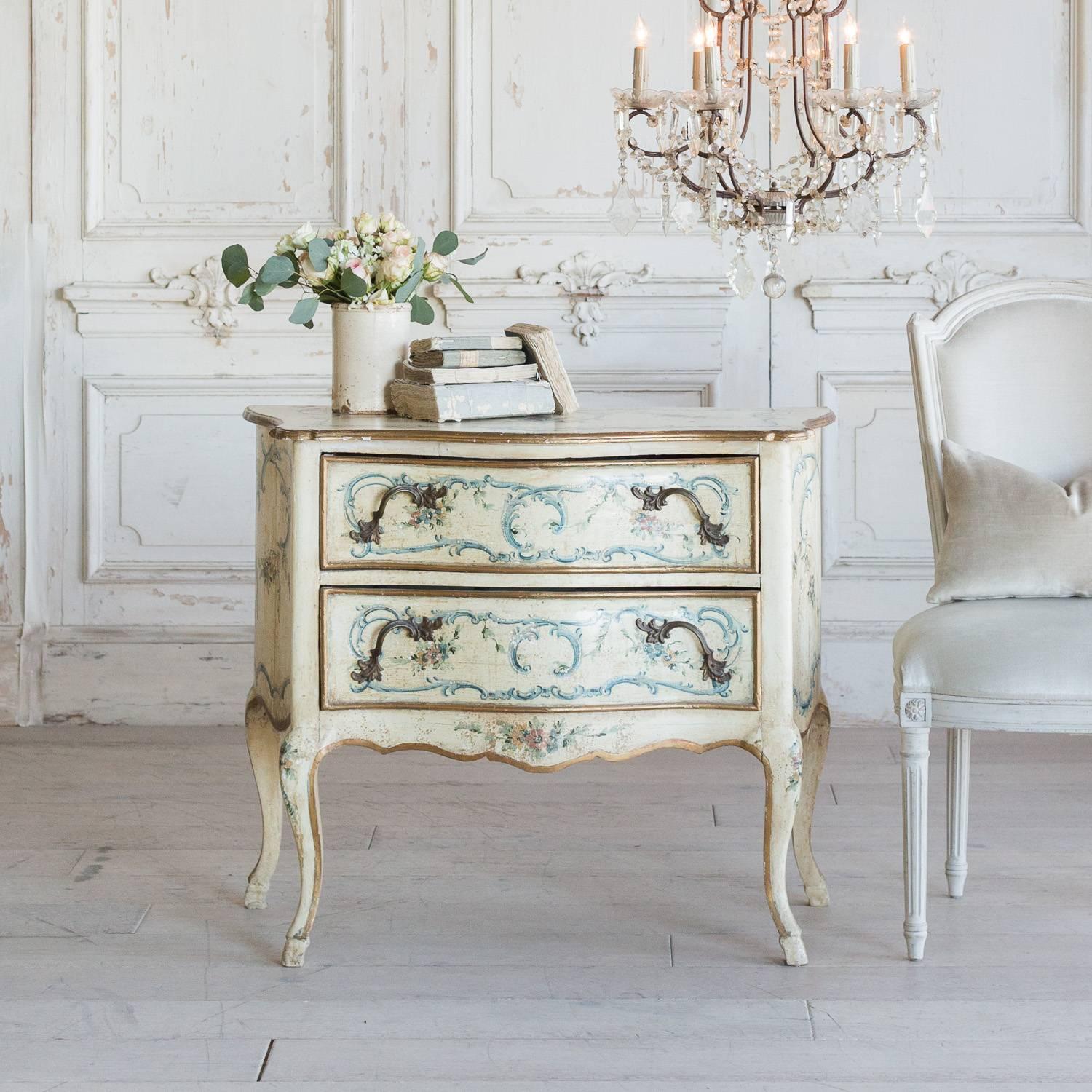 Charming antique nightstand commode in light buttercream finish with hand-painted flowers in pink and blue hues. Elaborate, Louis XV serpentine design adds charm and a touch of glamour to this piece. Two large drawers with a soft floral lining keep