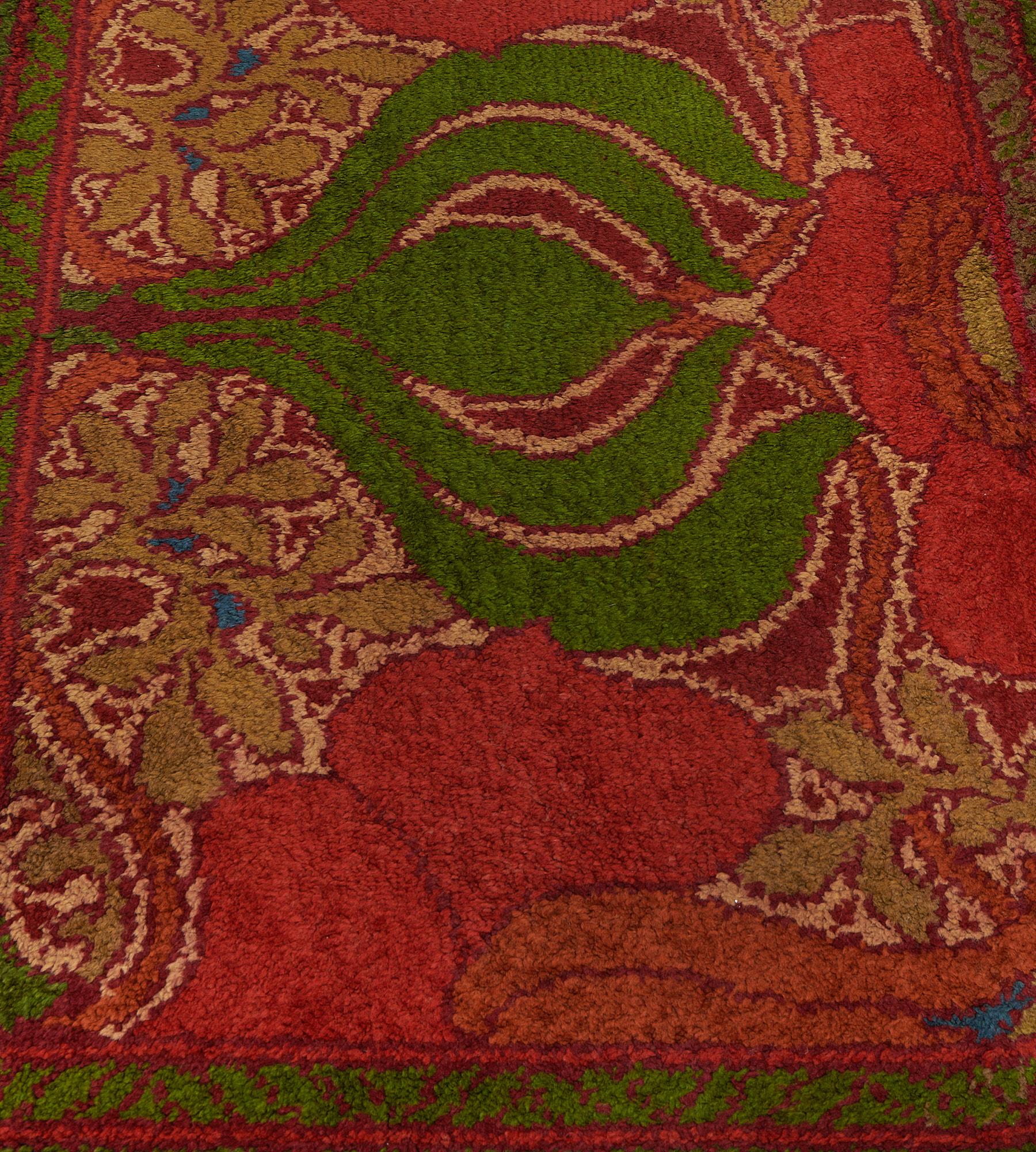 This antique, circa 1900, Irish Donegal Voysey rug features a field with a pair of large bottle-green onion shaped bulbs with a large alternatively facing tomato-red and orange large petal shaped flowerhead linked by moss-green and burgundy-red