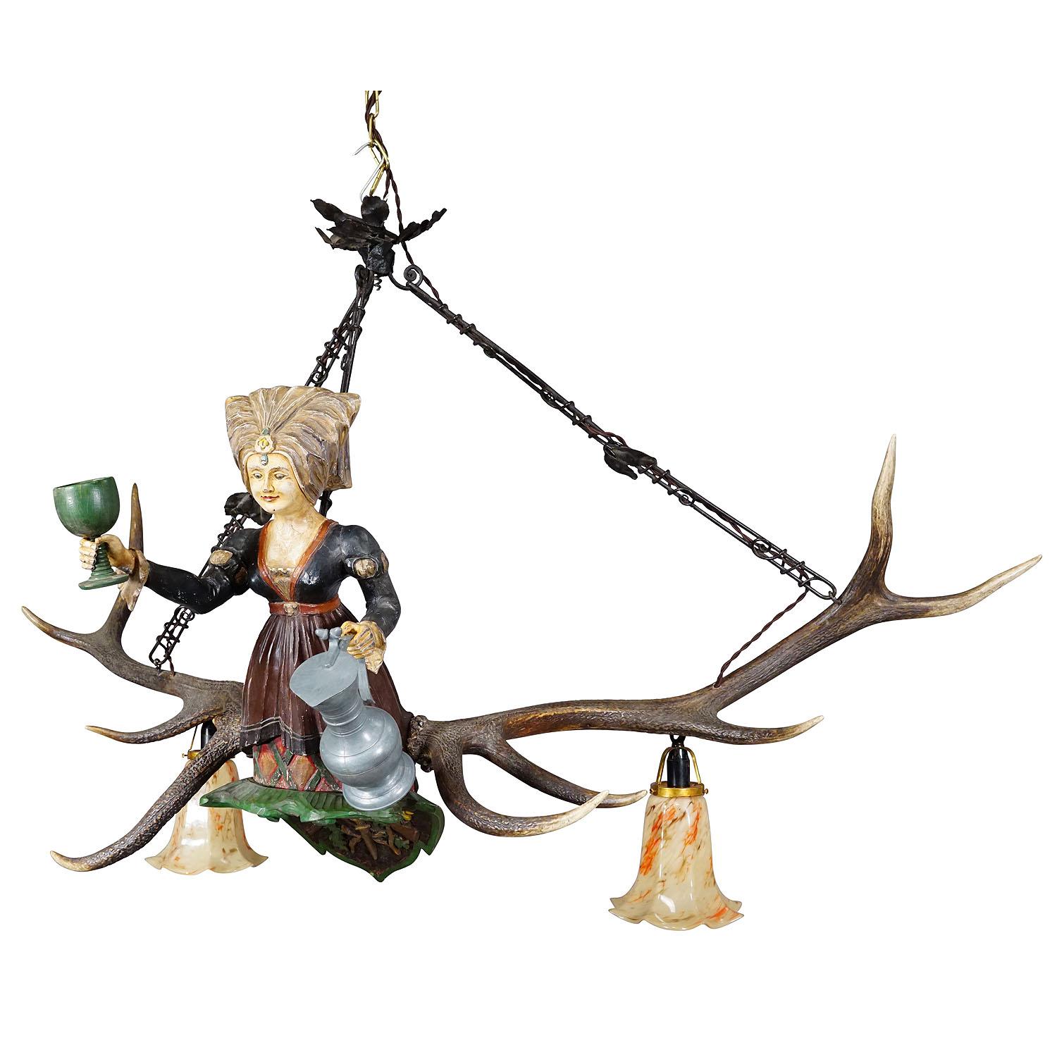 Antique Handcarved Lusterweibchen of a Wine Waitress ca. 1920

An antique lusterweibchen chandelier of a wine waitress lady holding a wine glass and a wine jug.  The luster is made of wood with handpainted finish and mounted on a large pair of deer