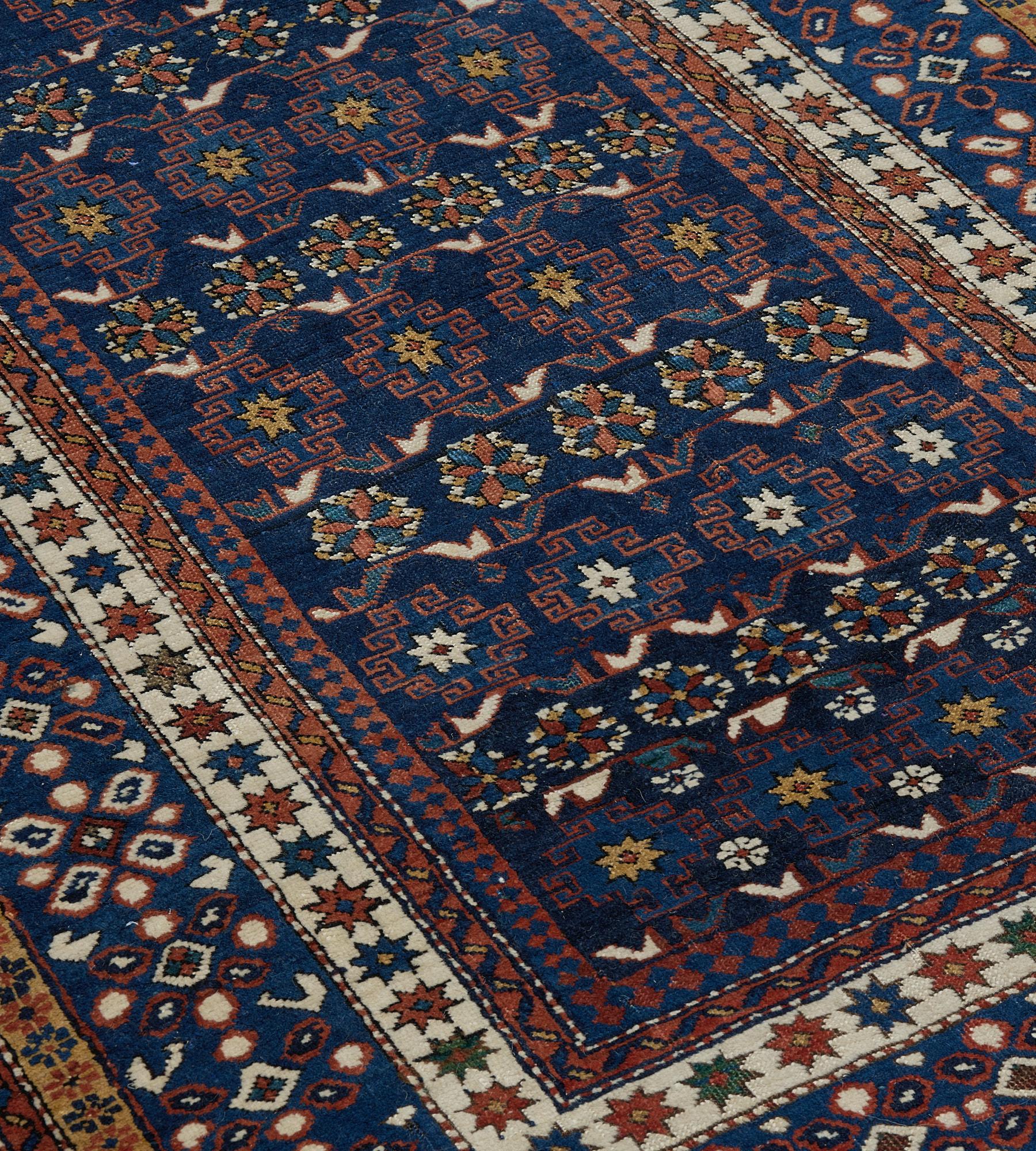 This antique, circa 1900, Chi-Chi rug has a shaded indigo-blue field with horizontal rows of light blue hooked linked lozenges between angular polychrome rosettes enclosed within a blue and terracotta-red reciprocal skittle-pattern stripe, in a