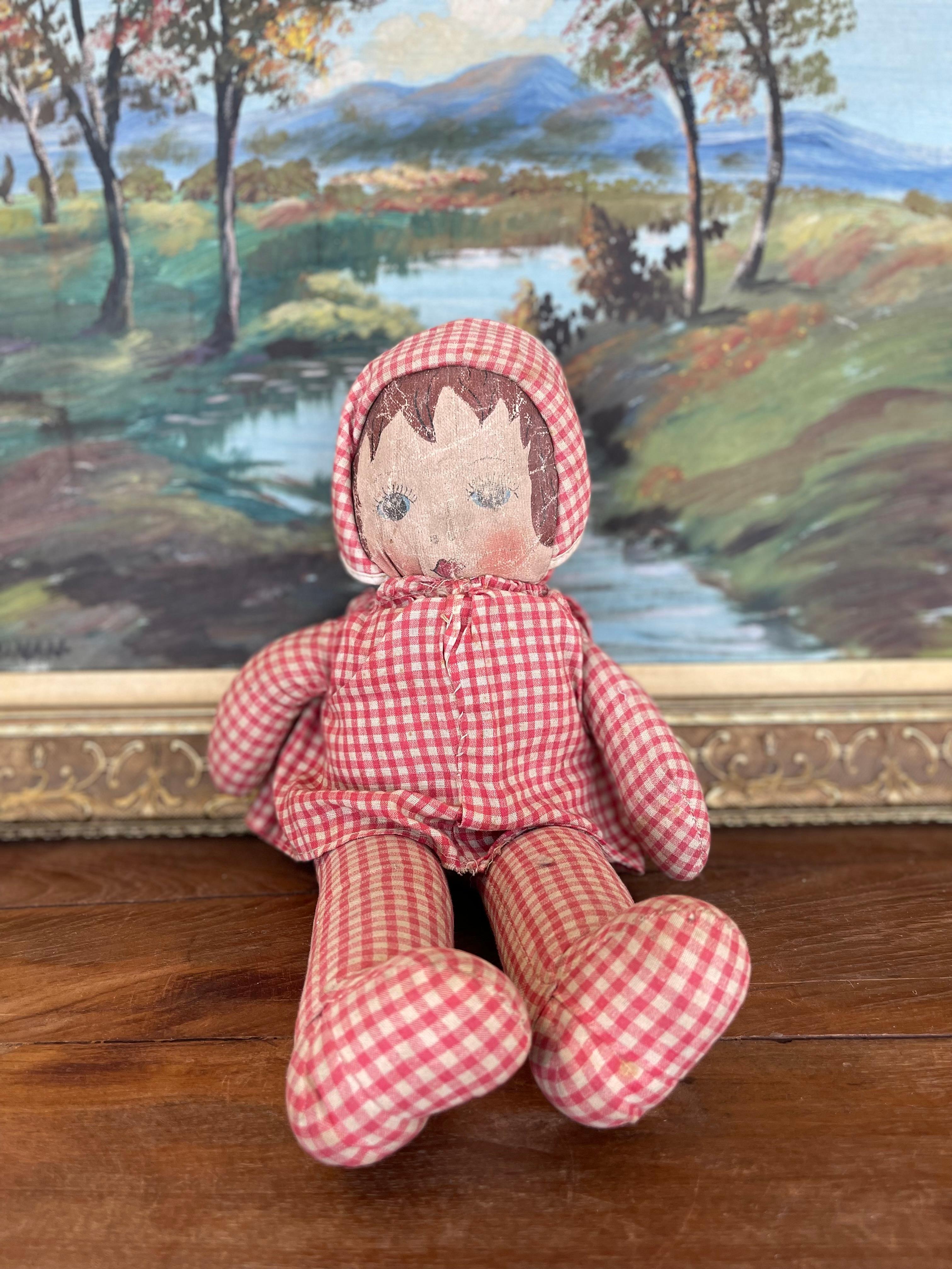 This antique handmade cloth doll dates to the early 20th century and features a charming hand-painted face with blue eyes, rosy cheeks, and short brown hair. The doll is dressed in its original red and white gingham outfit, complete with a
