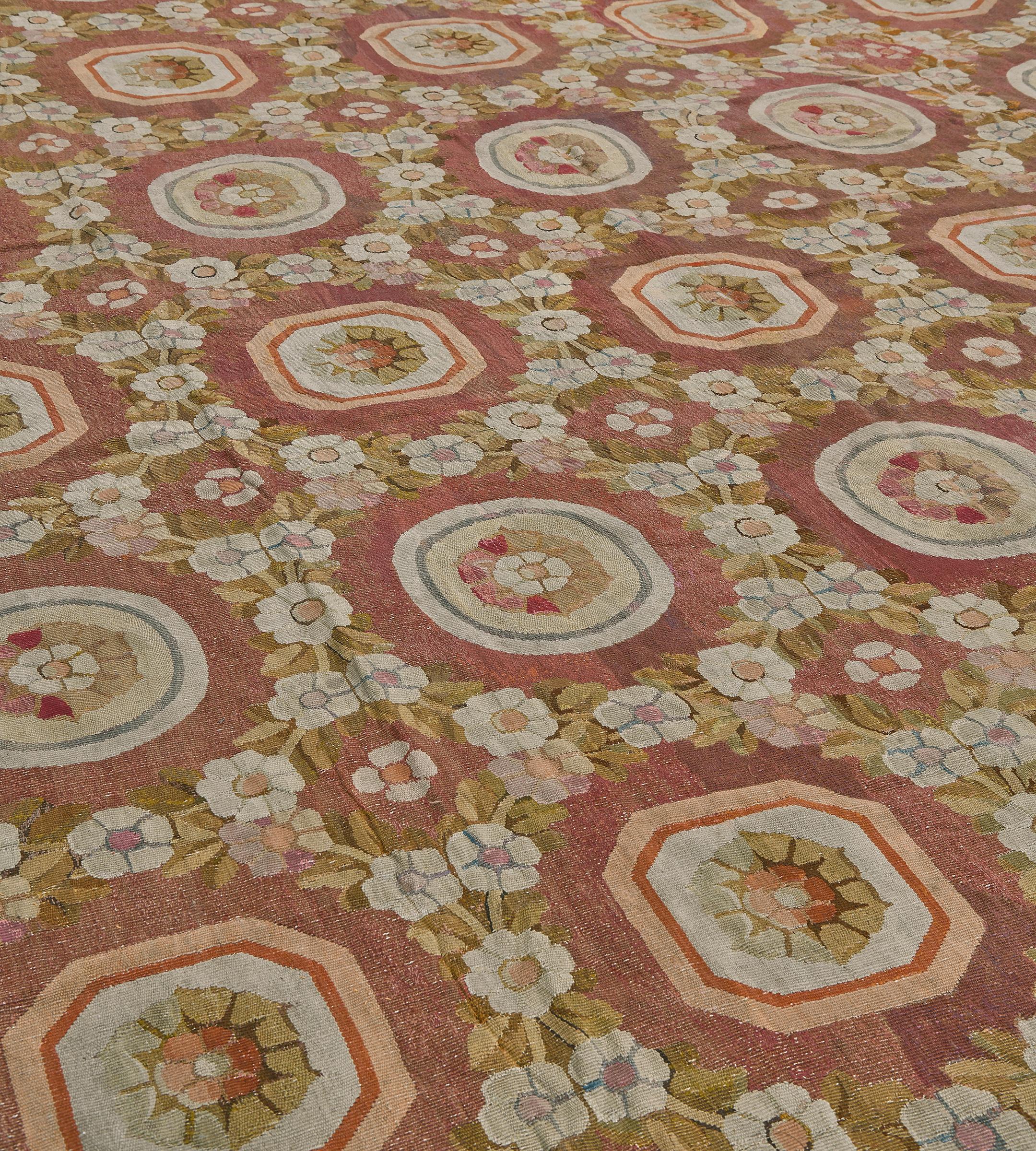 Questo antico tappeto Aubusson francese presenta un campo sfumato marrone-violaceo con una ghirlanda policroma di fiori e foglie intorno a file verticali di losanghe esagonali color avorio e giallo oro alternate a tondi contenenti ciascuno un