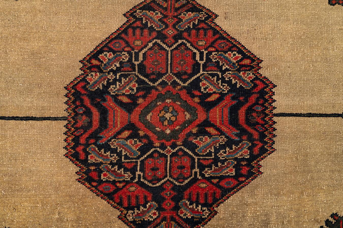 Dieser antike, handgewebte Malayer-Teppich zeigt ein auffälliges Zusammenspiel von kühner Geometrie und feiner Ornamentik. Das sanft gealterte sandbeige Feld bildet den Hintergrund für ein dramatisches Medaillon in der Mitte, das in tiefen Rot-,