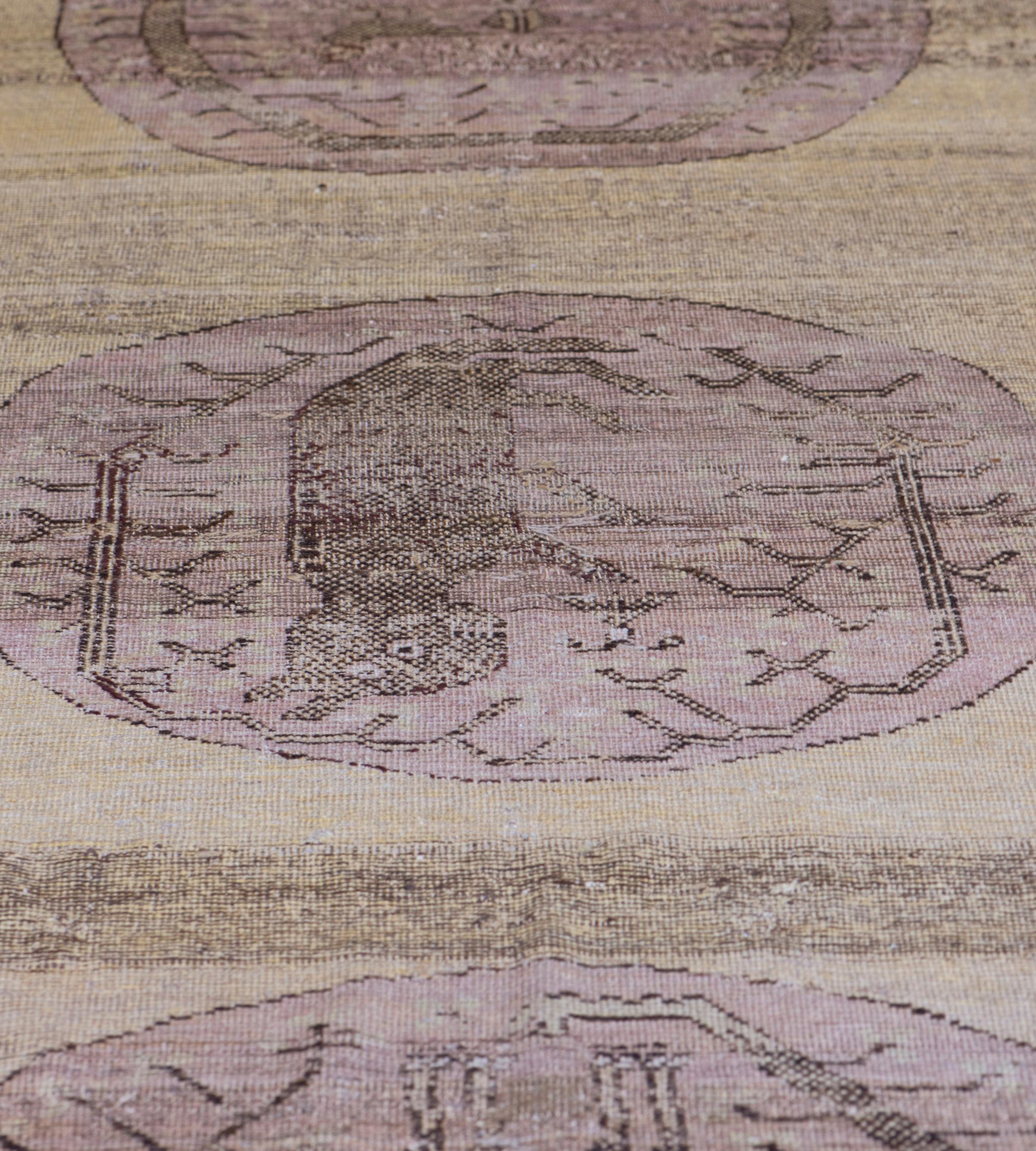 This antique Khotan has a shaded sandy-yellow field containing three large shaded purple roundels each containing an angular tree curving around various animals, in a narrow border of serrated leaves and cloud motifs between multiple flowerhead and