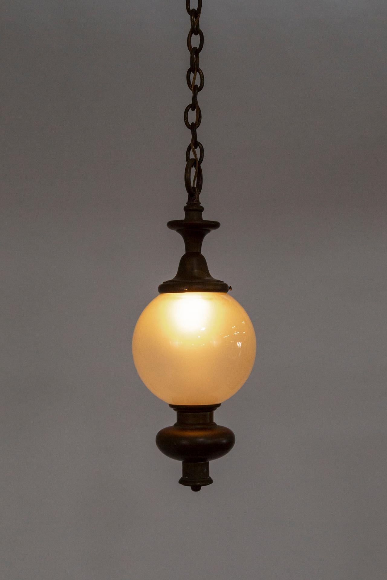 An early 20th-century pendant light with a frosted glass, globe shade adorned with a large, tiered, cylindrical, brass bottom finial and cap; hanging from a long chain with a matching canopy. The original blown glass shade gives off a lovely,
