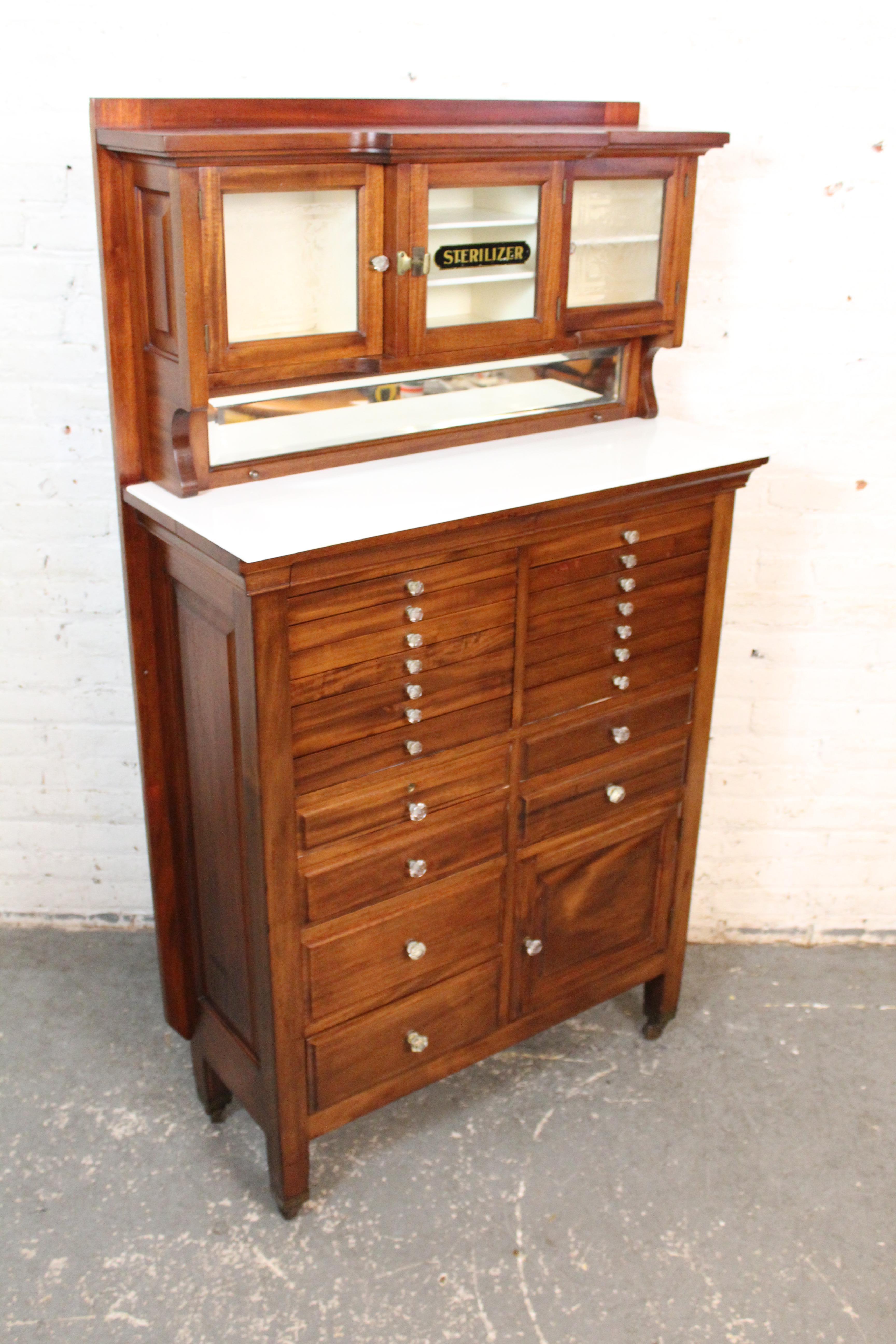 Antique dental cabinet made by The Harvard Company, Canton Ohio, featuring amazing details. Twenty two drawers and cabinet space below a glass front display case. In between sits a white glass top with beveled mirror that flips up and back for more