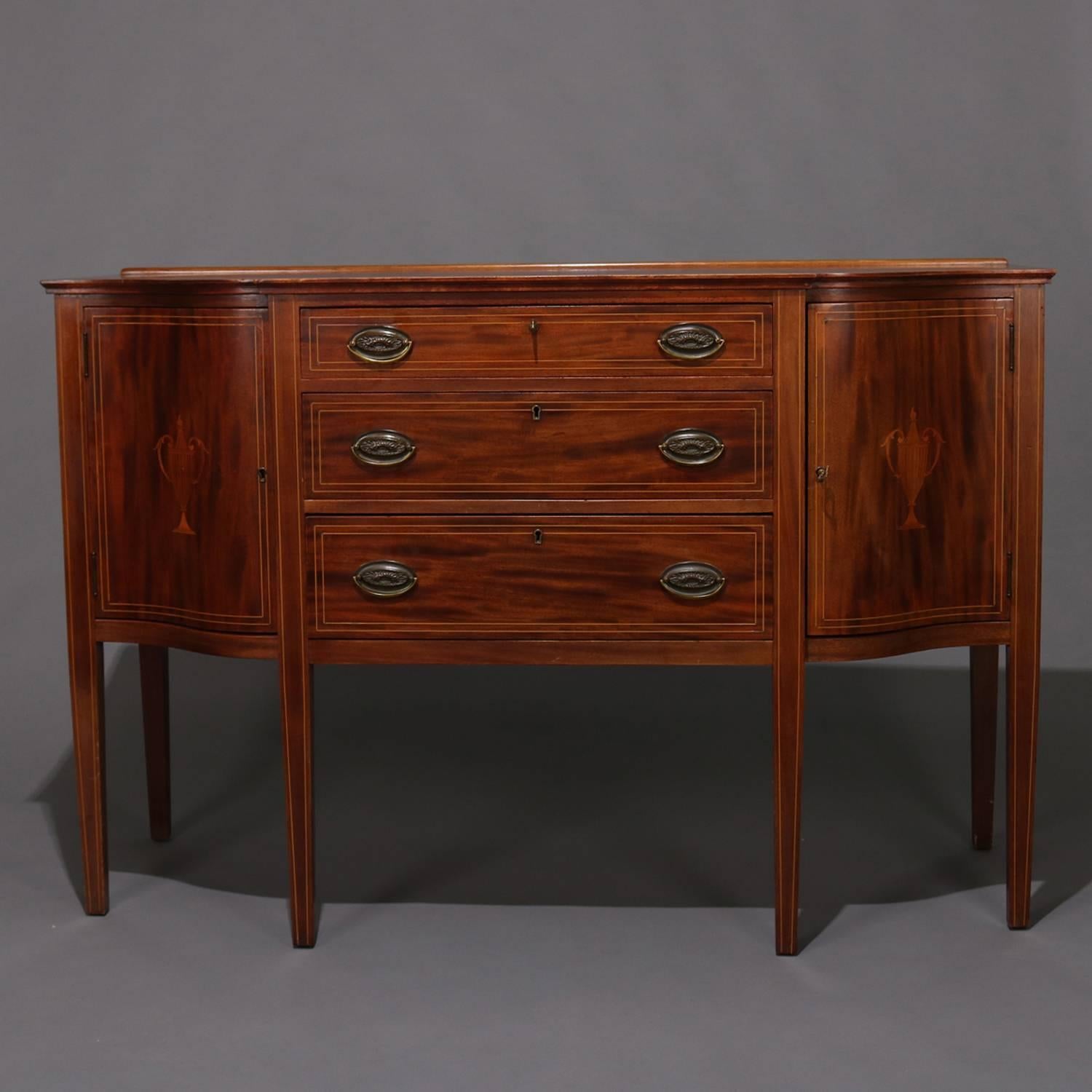 Antique Hepplewhite sideboard features deeply striated mahogany construction having three central drawers with flanking side cabinets and seated on tapered legs, satinwood banding throughout, inlaid side doors having urn motif, bronze pulls, locking