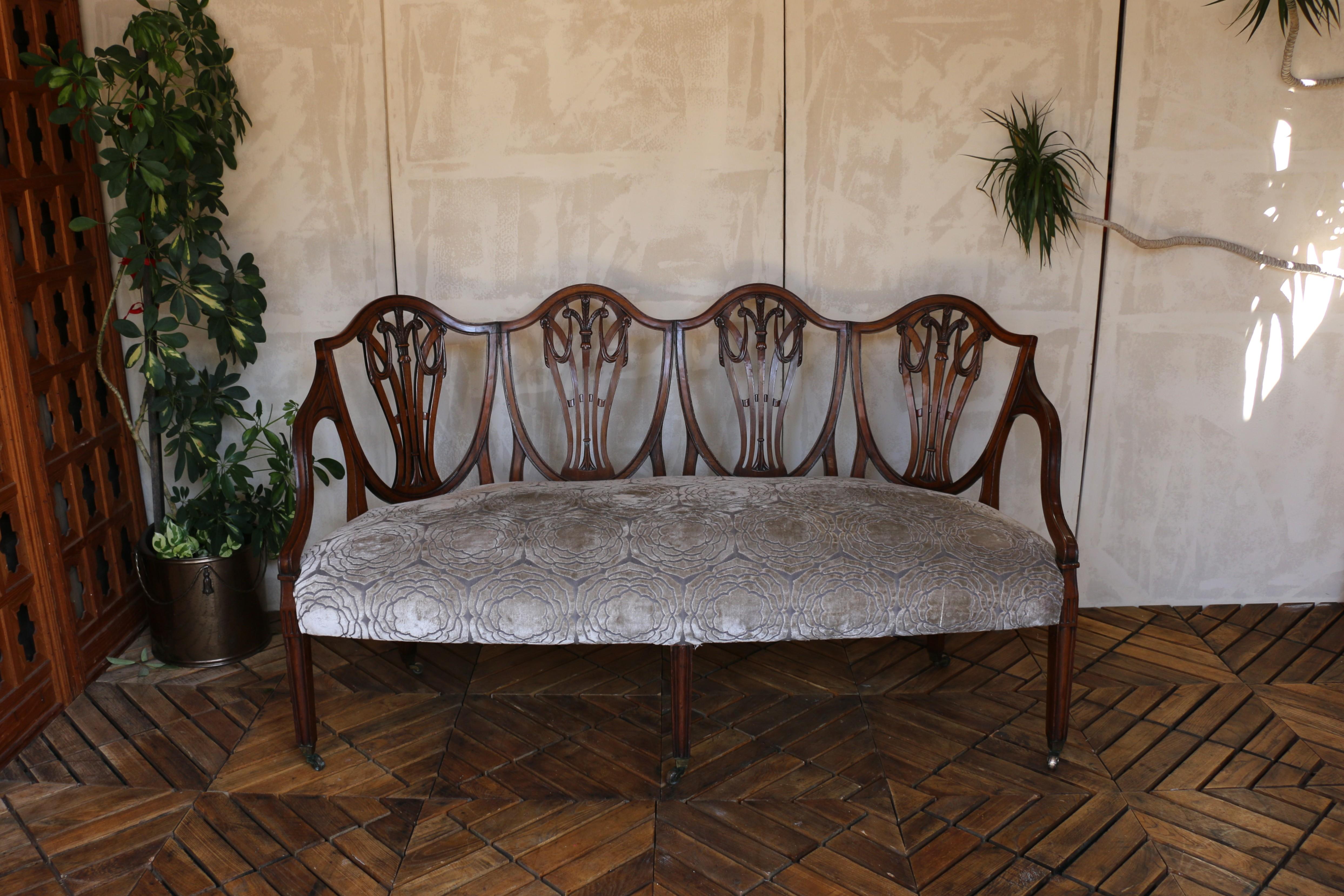 This elegant Hepplewhite-style settee dates to the late 19th century, circa 1890s England. Expertly crafted from solid mahogany, it features the distinctive quadruple shield-back design, each section finely carved with graceful motifs typical of the
