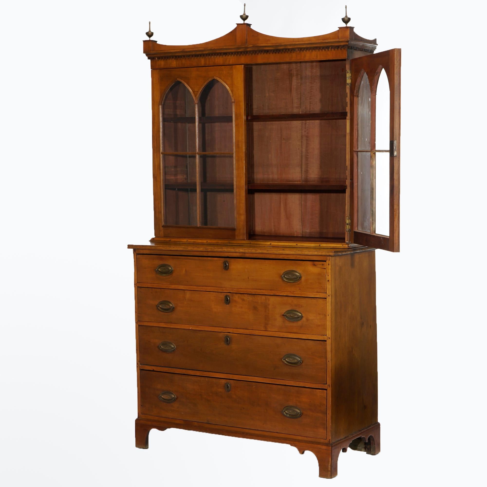 An antique English Hepplewhite breakfront cabinet offers cherry construction with shaped crest having three turned finials over cabinet with double doors with arced glass panels and opening to shelved interior over lower case with four graduated