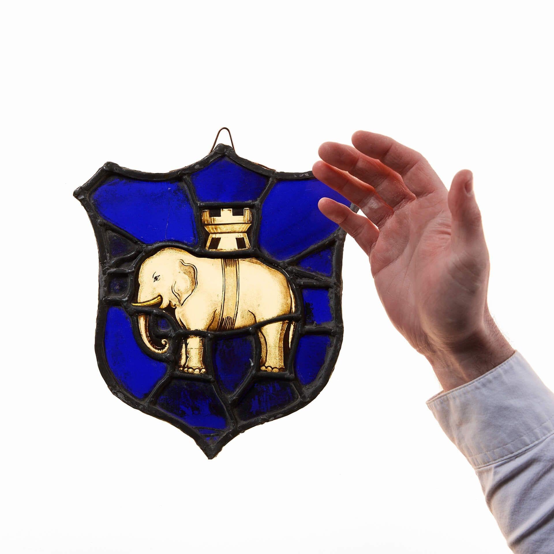 Eine Bleiverglasung im heraldischen Stil des späten 19. Jahrhunderts mit einem Wappen auf einer Schildform.

Dieser kleine Glasbehang bringt mit einem handgemalten Elefanten und einer Burgspitze auf leuchtend blauem Hintergrund ein Element der