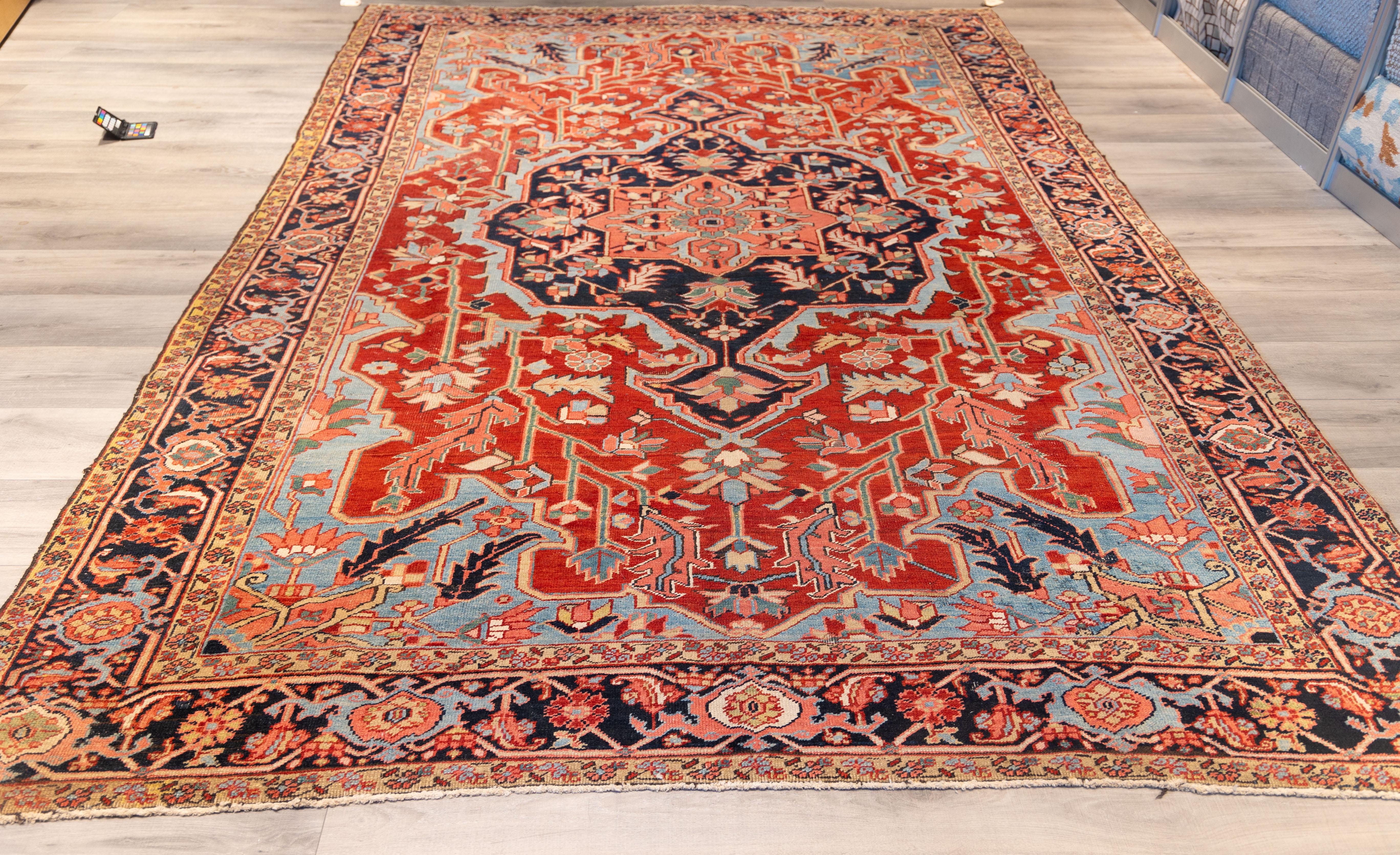 finely woven Heriz carpet. A dramatic midnight-blue medallion with classic angular symmetry, set against a richly rust-red field bursting with stylized foliage and serrated leaves. Pale sky-blue corners. 
The composition is framed by a wide border