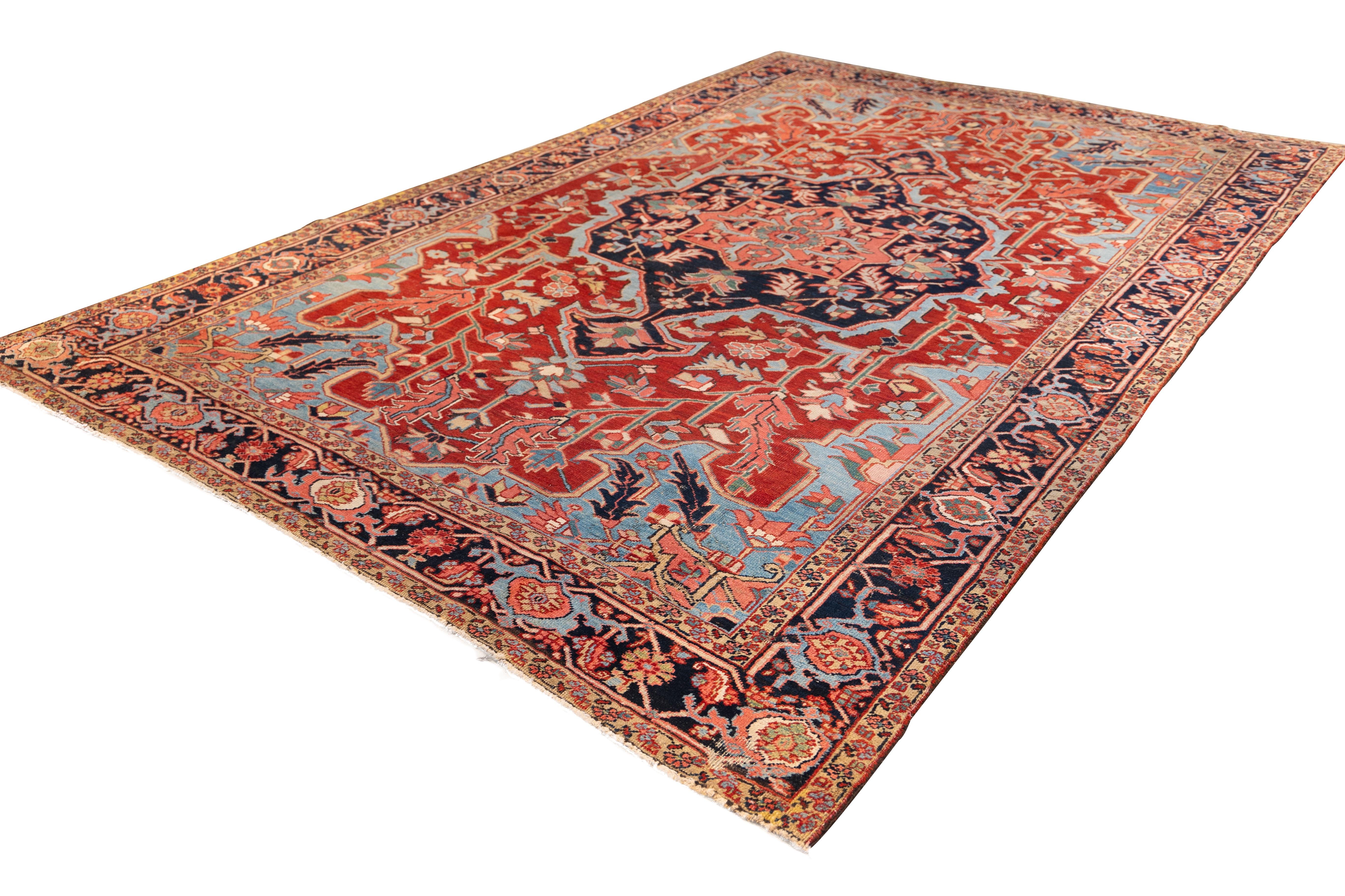 finely woven Heriz carpet. A dramatic midnight-blue medallion with classic angular symmetry, set against a richly rust-red field bursting with stylized foliage and serrated leaves. Pale sky-blue corners. 
The composition is framed by a wide border