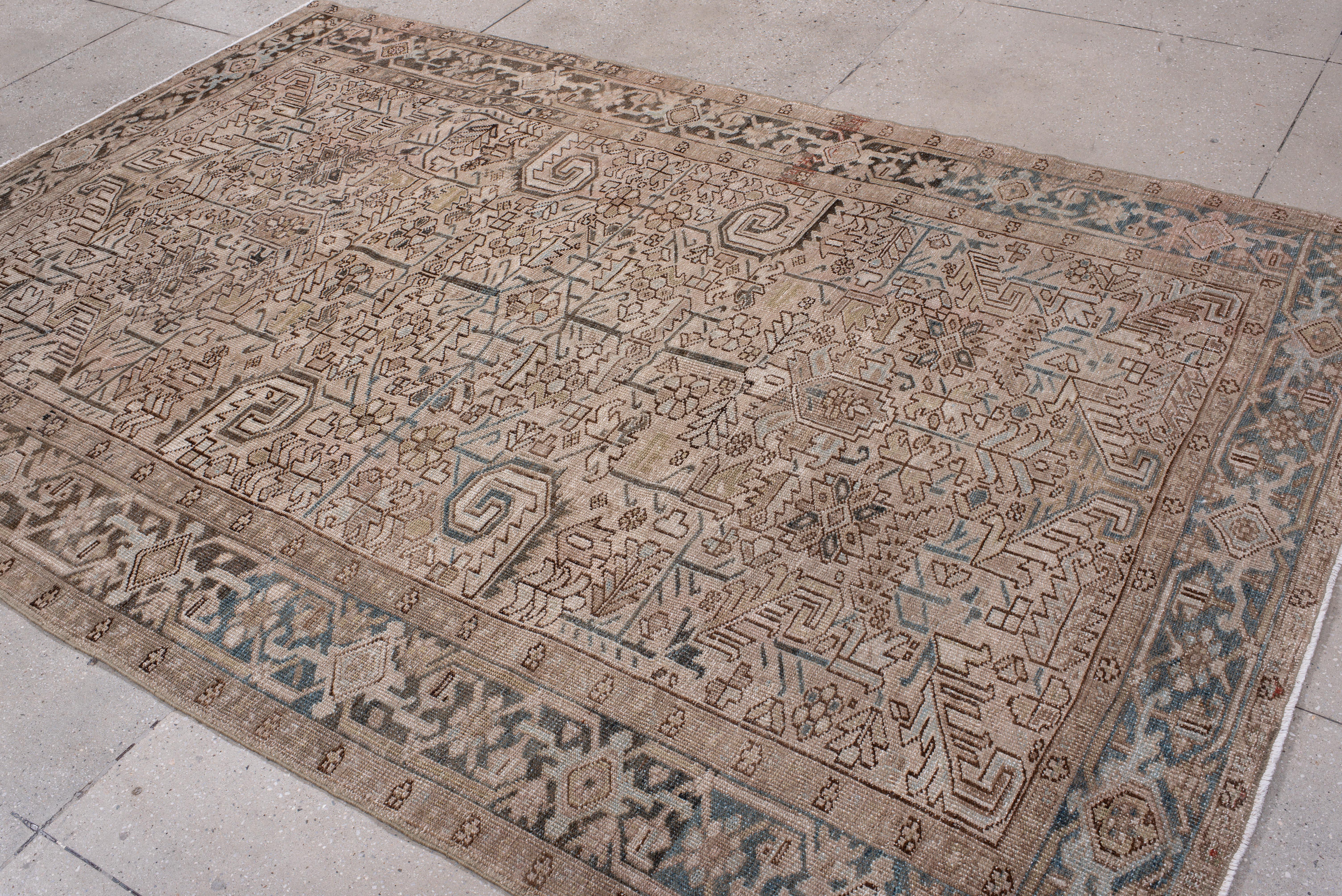 This antique Heriz rug is defined by a powerful geometric medallion structure woven in a palette of taupe, sand, cocoa, and pale slate blue. Its central layout bursts with angular palmettes, serrated leaves, and stylized rosettes, bordered by a