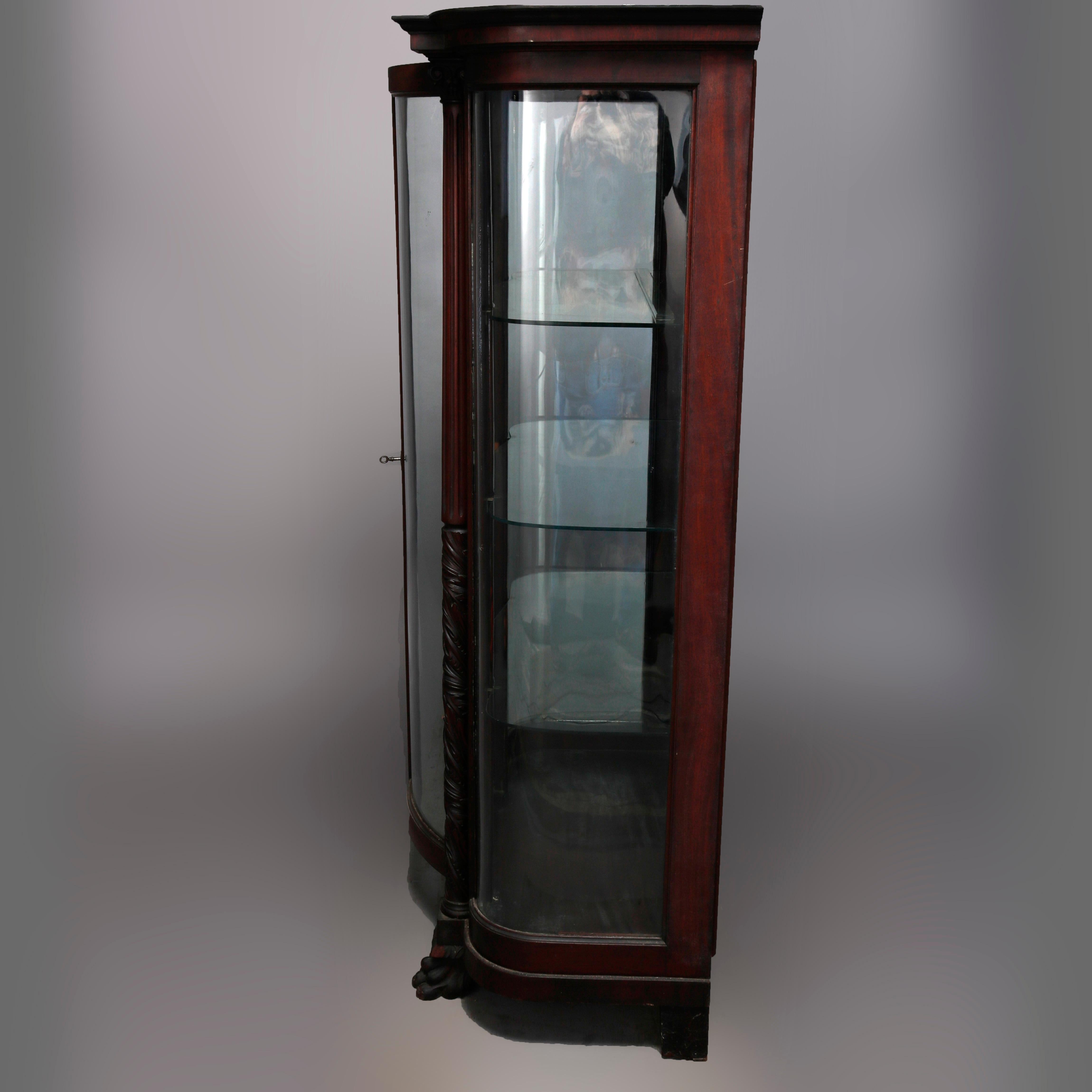 An antique American Empire china cabinet by Hersee & Co, Buffalo, New York offers mahogany construction in with bowed cabinet in diminutive form having curved glass sides and single door opening to shelved interior and flanked by carved Corinthian