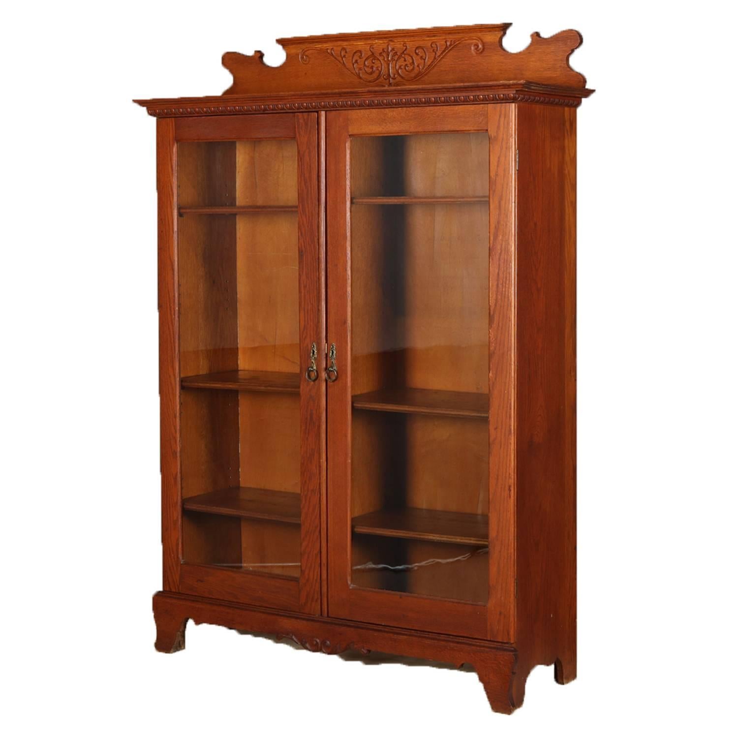 Antique Horner Bros. School oak bookcase features shaped backsplash with incised scroll and foliate motif above case with egg and dart ti-rimming and double glass doors opening to adjustable shelf interior, having shaped skirt flanked by bracket
