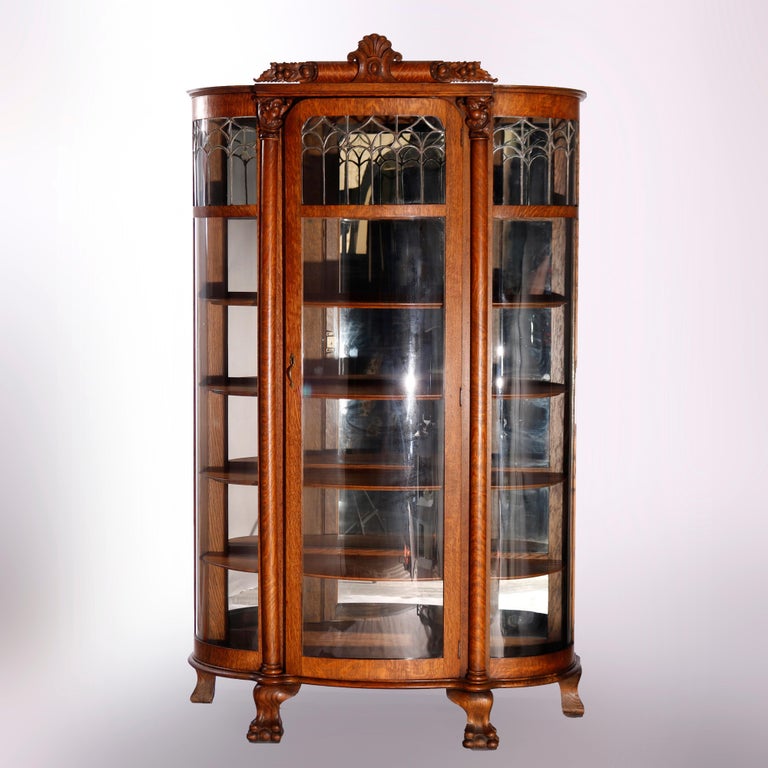 An antique Horner bow-front china cabinet offers quarter sawn oak construction with crest having central carved shield cartouche over single door mirrored cabinet having curved glass with leaded decoration, shelved interior, flanking supports with
