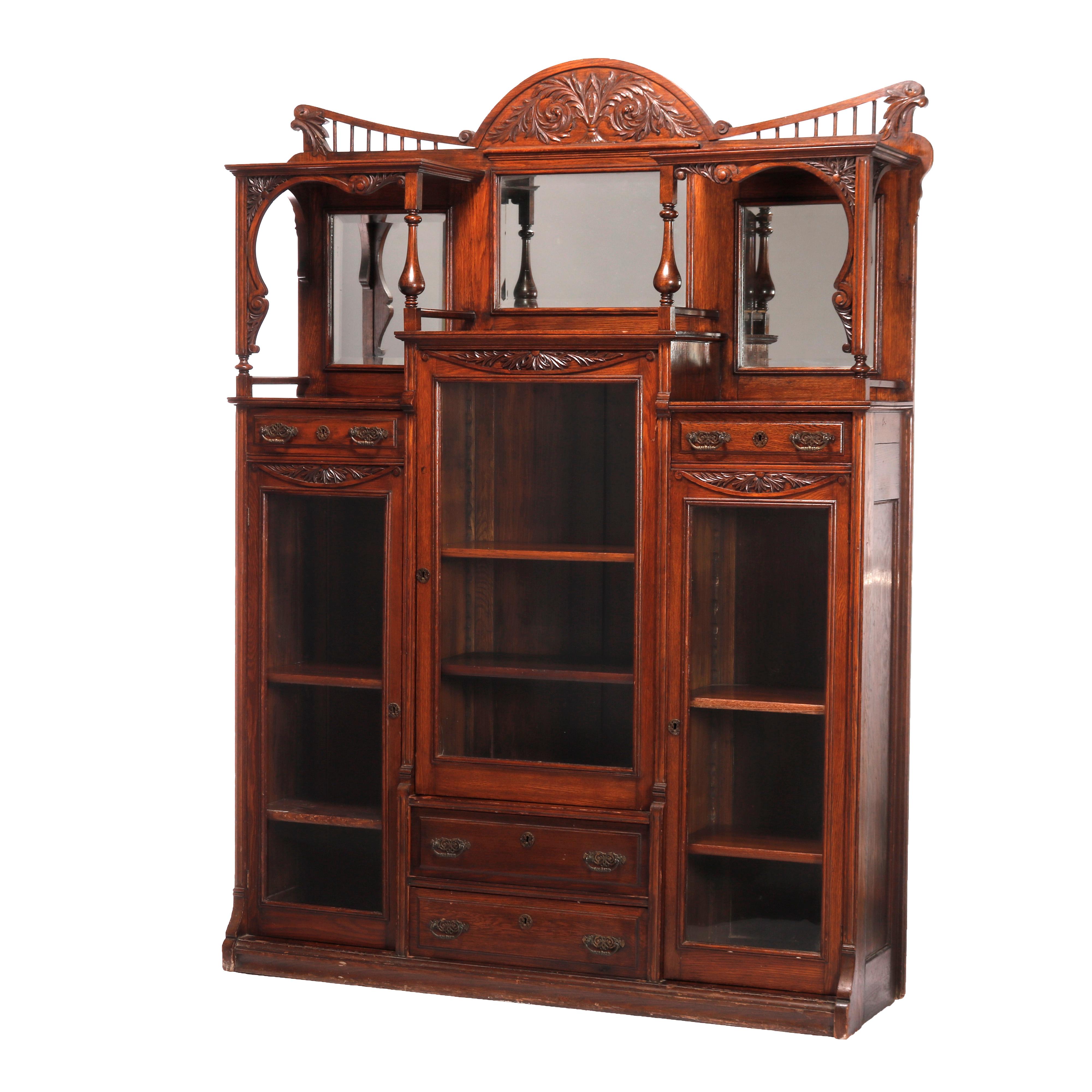 An antique side-by-side bookcase in the manner of RJ Horner offers quarter sawn oak construction with crest having carved palmette floral design and flanking spindle gallery surmounting triple case with mirrored backsplash over towers having drawers
