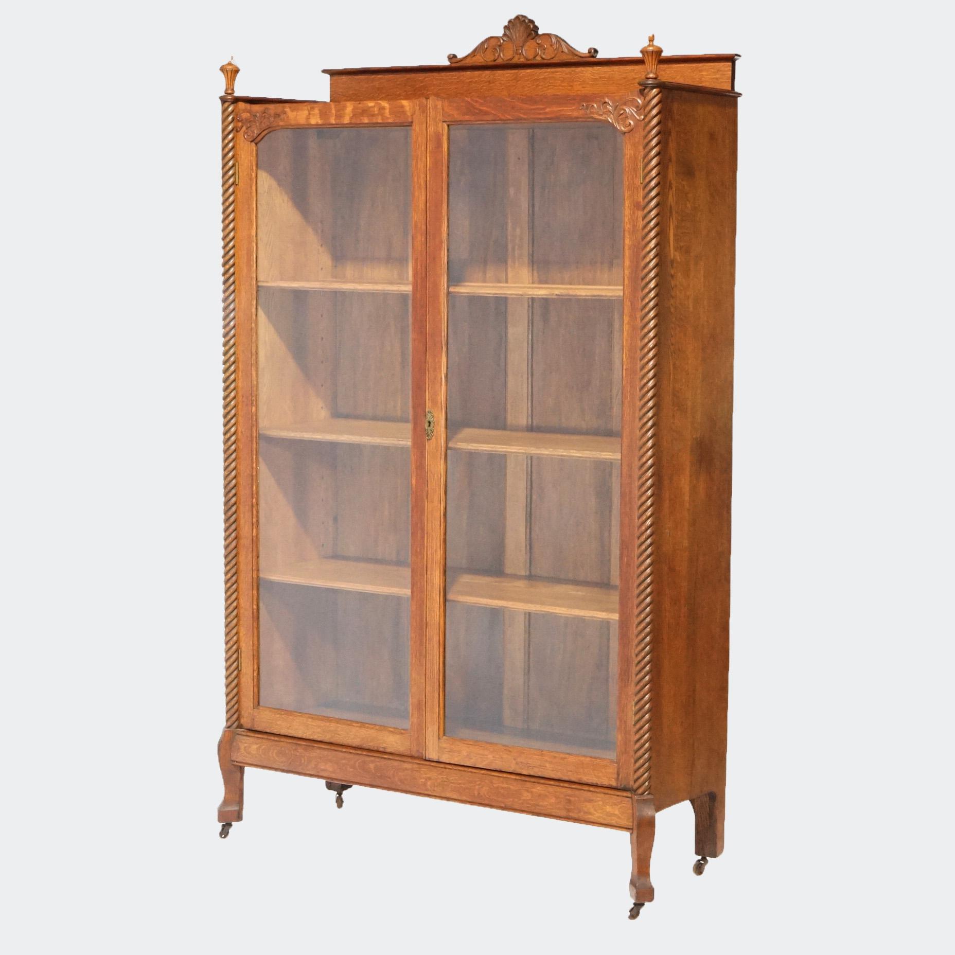 An antique bookcase in the manner of RJ Horner offers oak construction with foliate carved crest over double door case having flanking rope twist supports and shelved interior, raised on stylized cabriole legs terminating in spade feet, circa