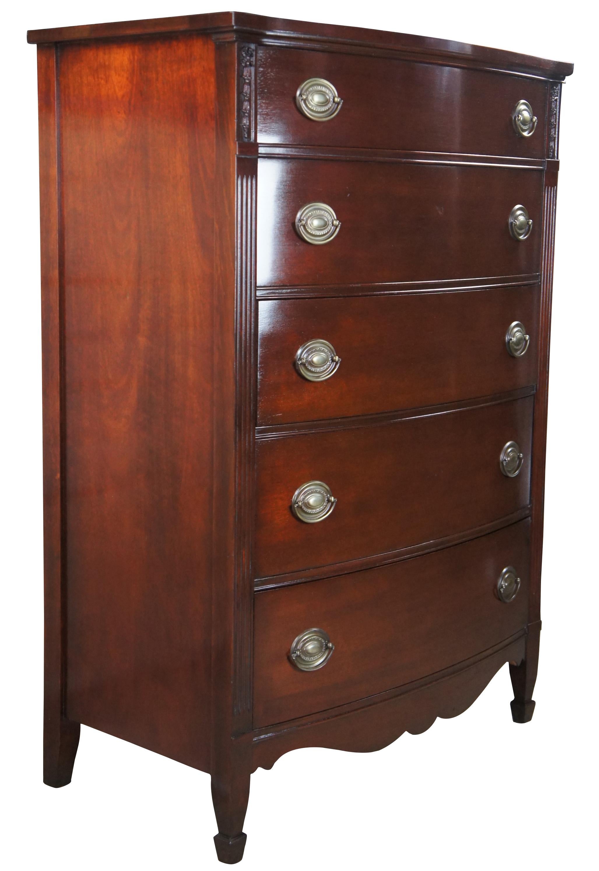 A beautiful Sheraton style tallboy dresser by Huntley Furniture, circa 1930s. Made from mahogany with a bow front. Includes 5 dovetailed drawers, oval brass hardware, carved foliate, and fluted stiles along the sides. The chest is supported by