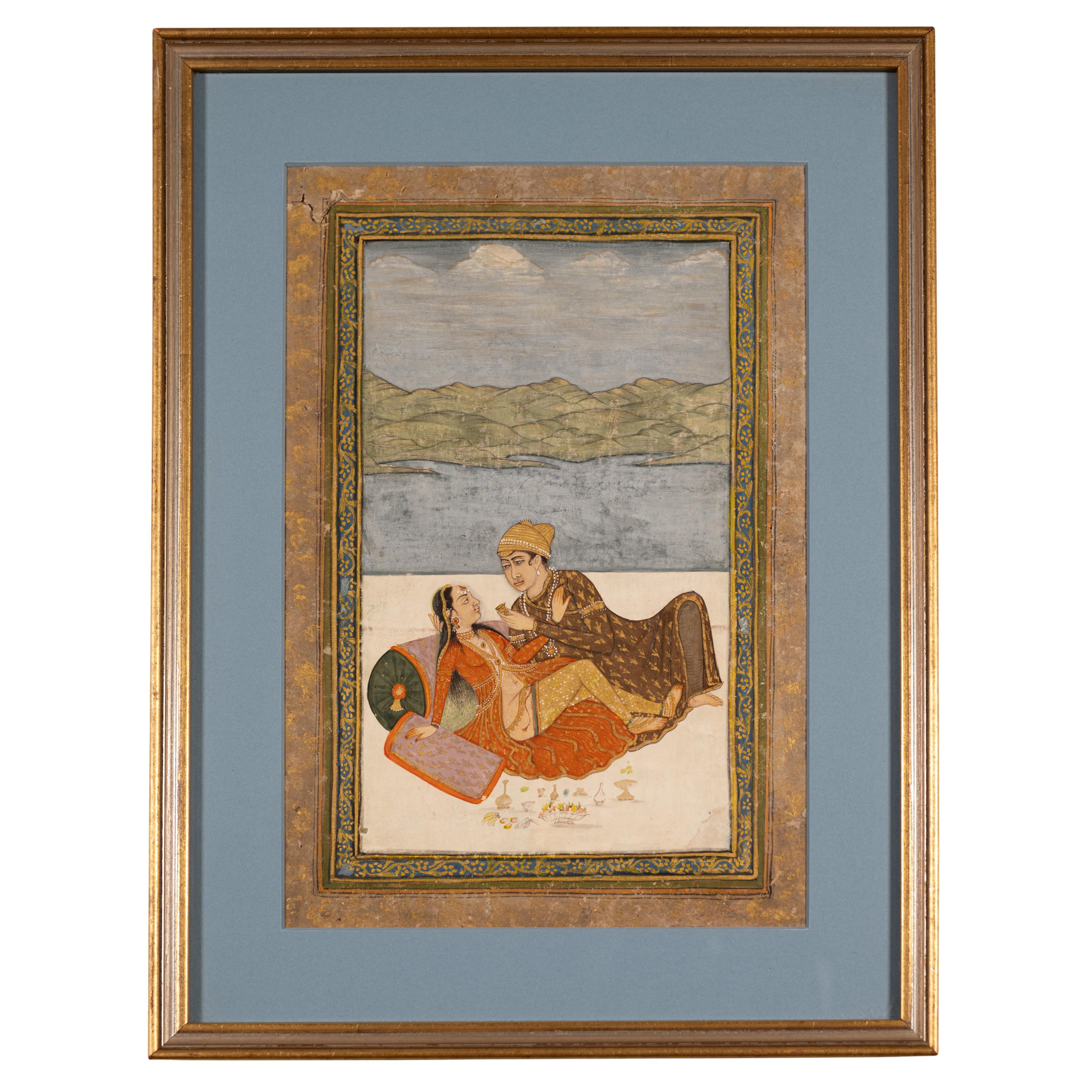Offered here is a gouache and gold miniature painting on paper of an amorous couple on a veranda set against a mountain and lake background.
The male dressed in a princely attire is leaning over his semi-clad lover resting against a bolster. He is