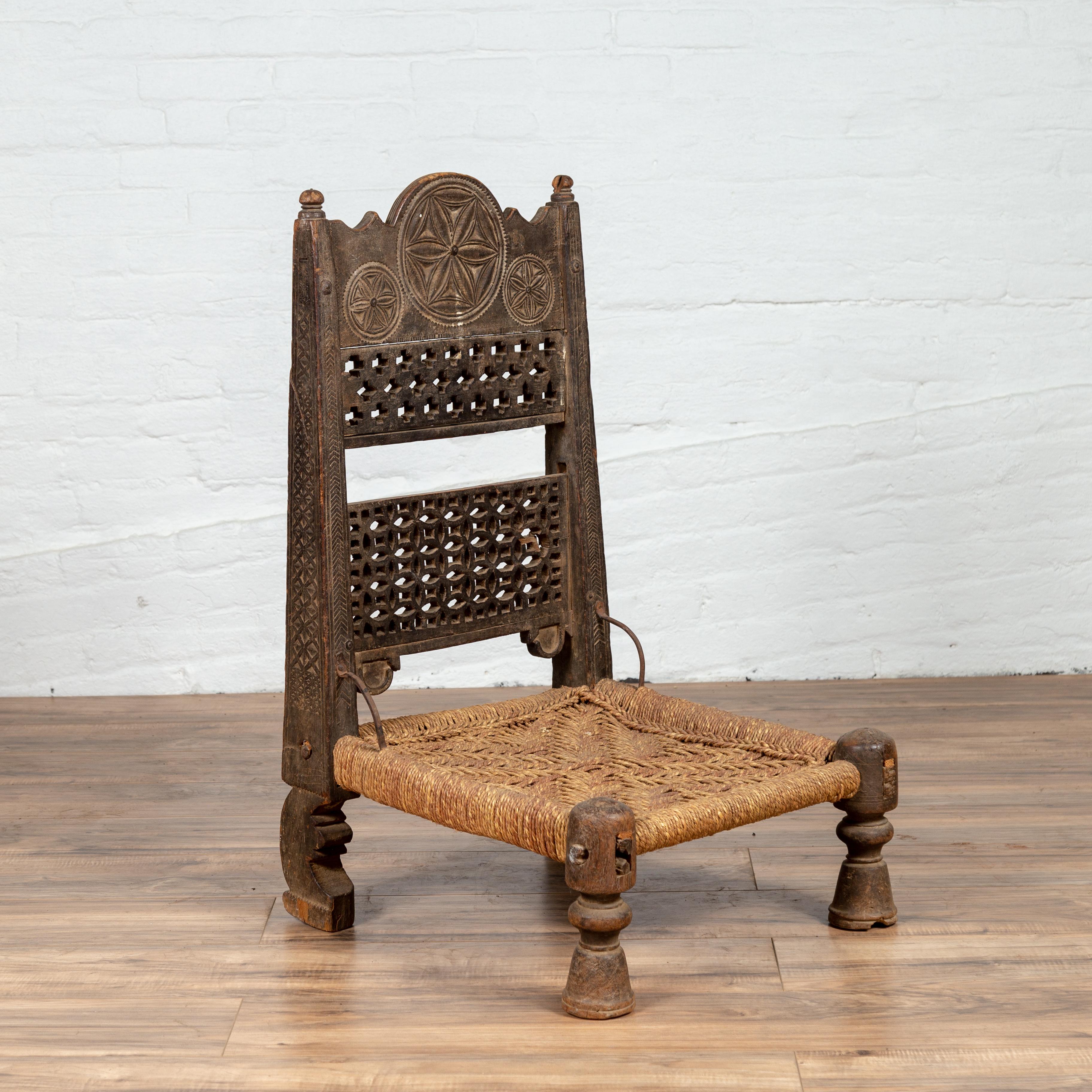 An antique Indian rustic low wooden chair from the 19th century, with fretwork accents, carved rosette and handwoven twine. Charming our eyes with its rustic appearance, this low wooden chair features a straight back, adorned with rosettes hand