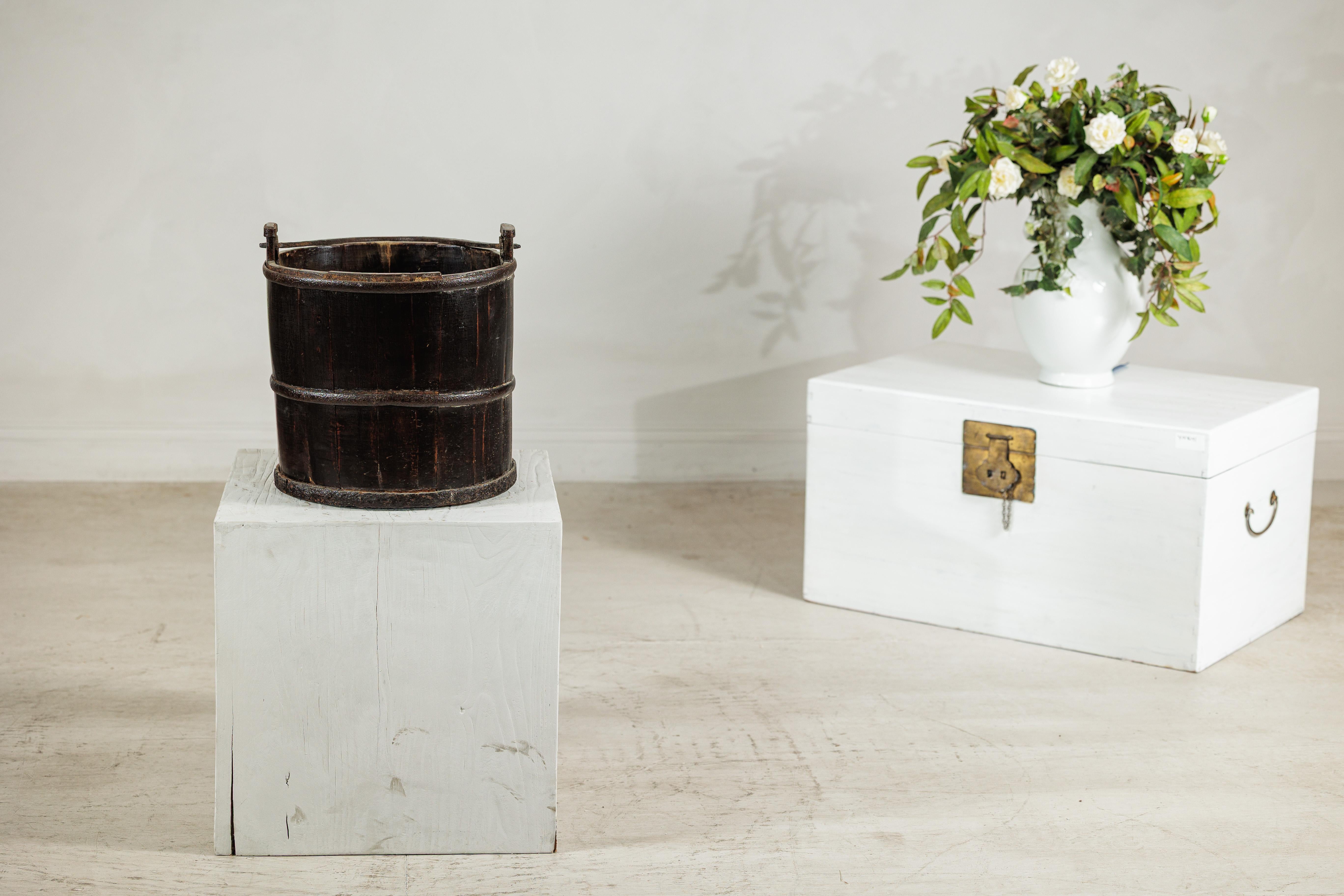 An evocative antique Indian wooden water bucket, handcrafted from dark-stained timber and bound with iron bands. The bucket’s cylindrical form is composed of vertical staves joined by two broad metal hoops, showing traces of age, wear, and use that