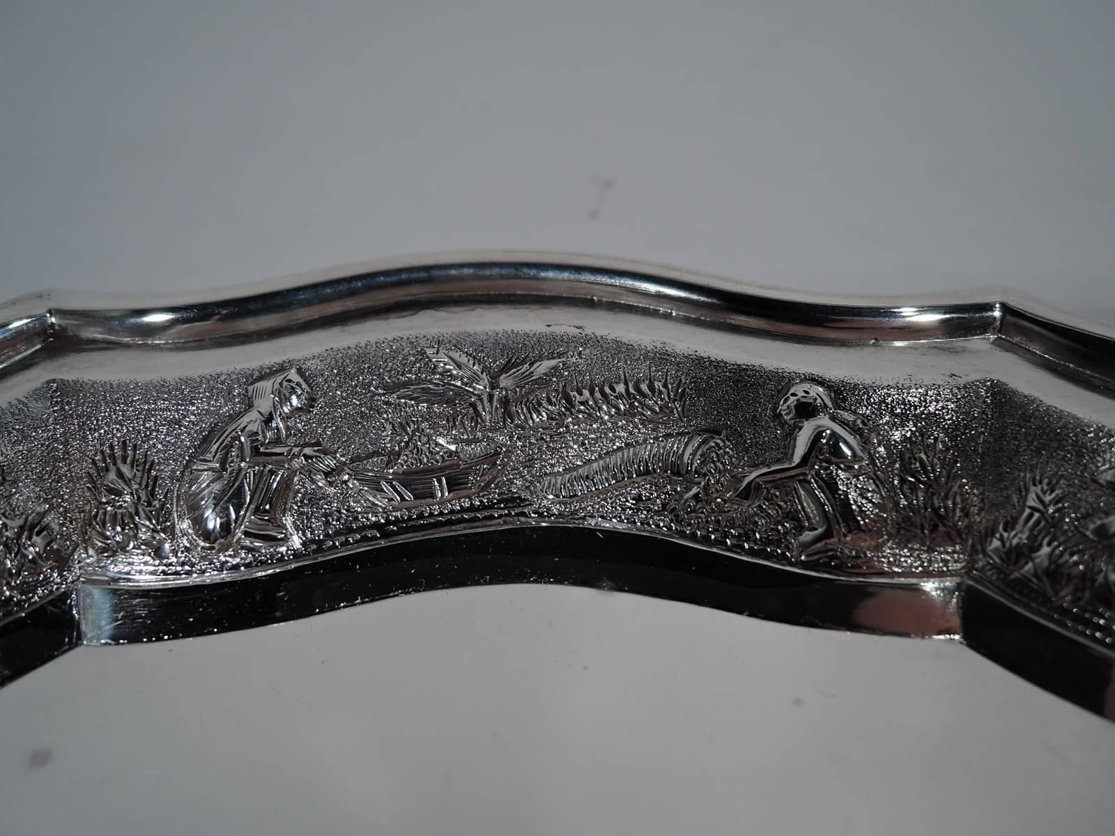 Antique Indo-Chinese silver tray, circa 1910. Circular with curvilinear piecrust rim. Tapering sides with rural scenes in low relief on stippled ground: Farmers, hunters, thatched buildings, and flourishing vegetation. An unusual mix of Eastern