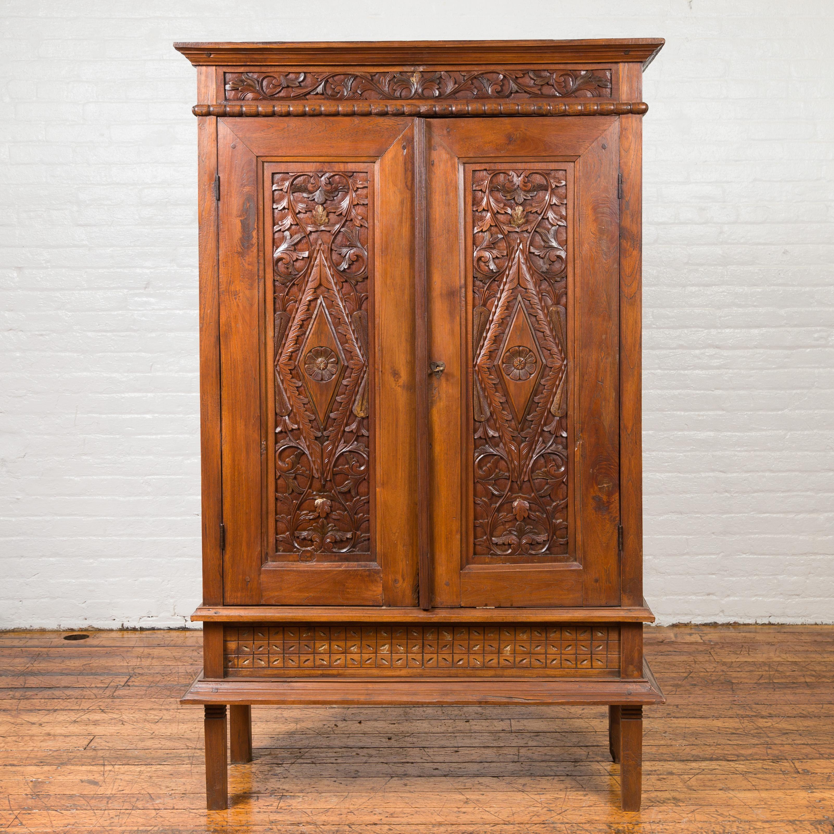 An antique Indonesian wooden cabinet from the 19th century with carved scrolling foliage. Capturing our attention with its clean lines and richly carved décor, this Indonesian cabinet features a molded cornice sitting above a frieze of scrolling