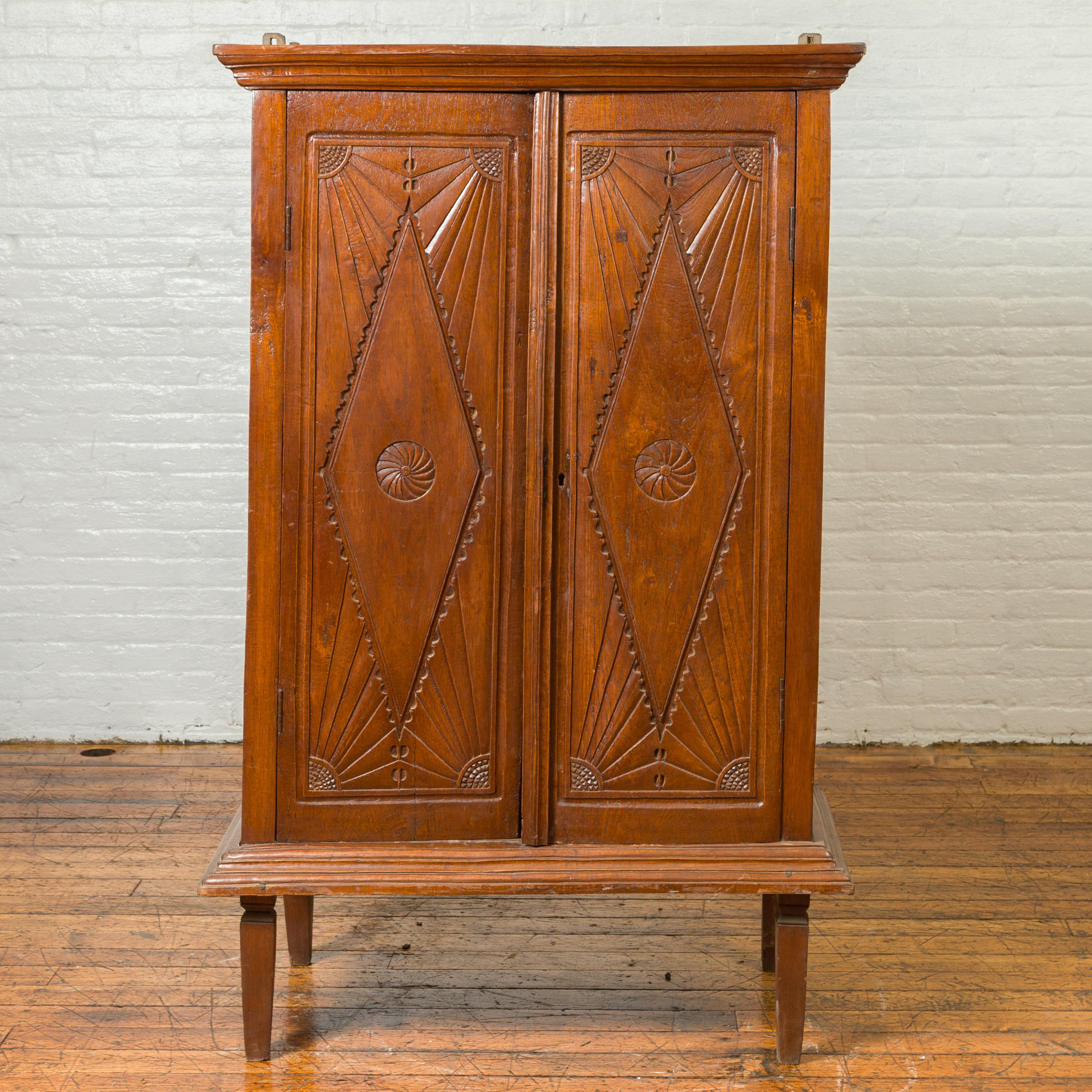 A Dutch Colonial wooden cabinet from the 19th century, with carved doors, diamonds and radiating motifs. Immerse yourself in the historical depth and artistic beauty of this 19th-century Dutch Colonial wooden cabinet, an artifact that harmoniously