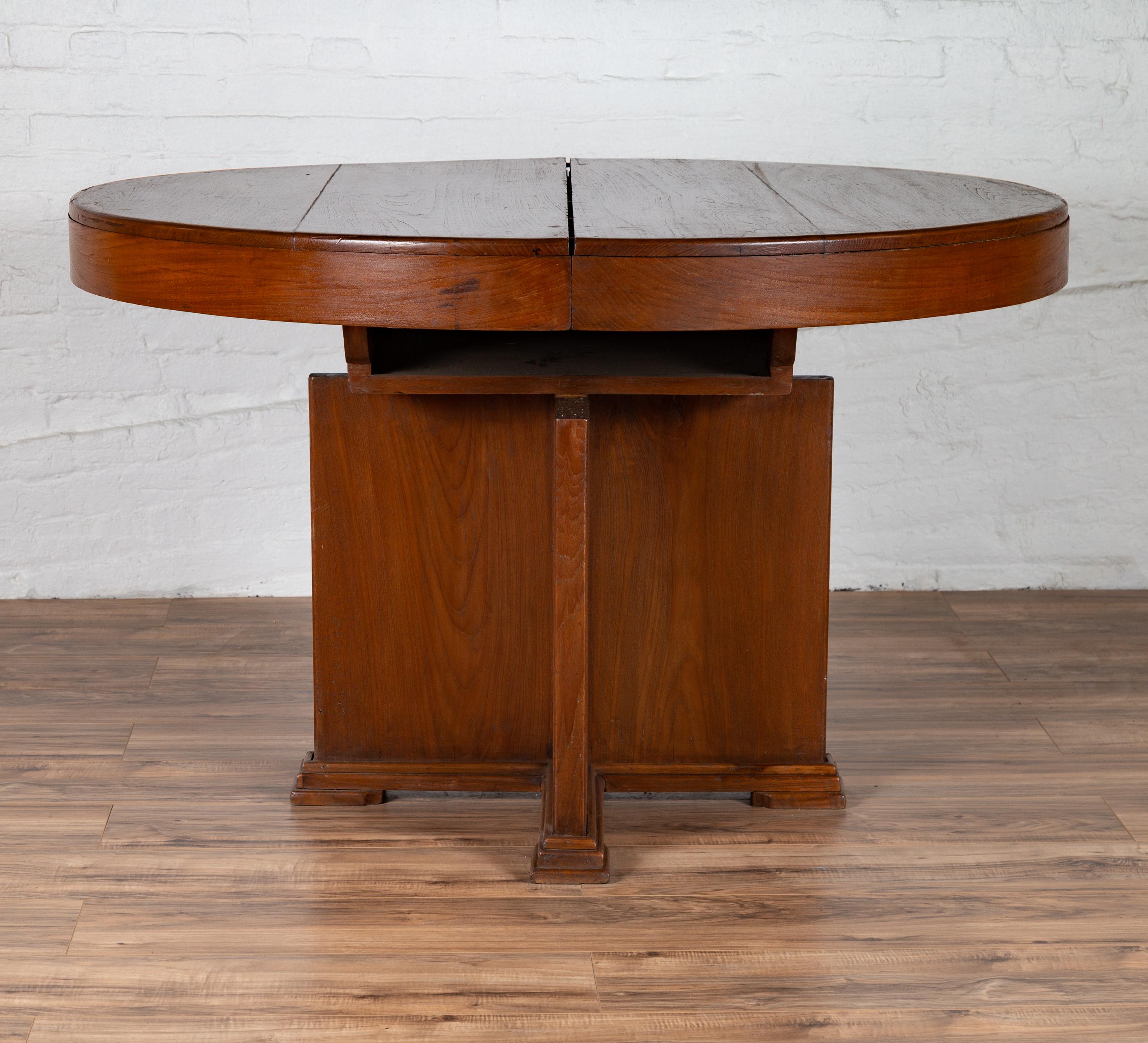 An antique Indonesian wooden dining table from the early 20th century, with central folding leaf and geometric base. Born in Indonesia during the early years of the 20th century, this stylish dining table features a circular planked top concealing a