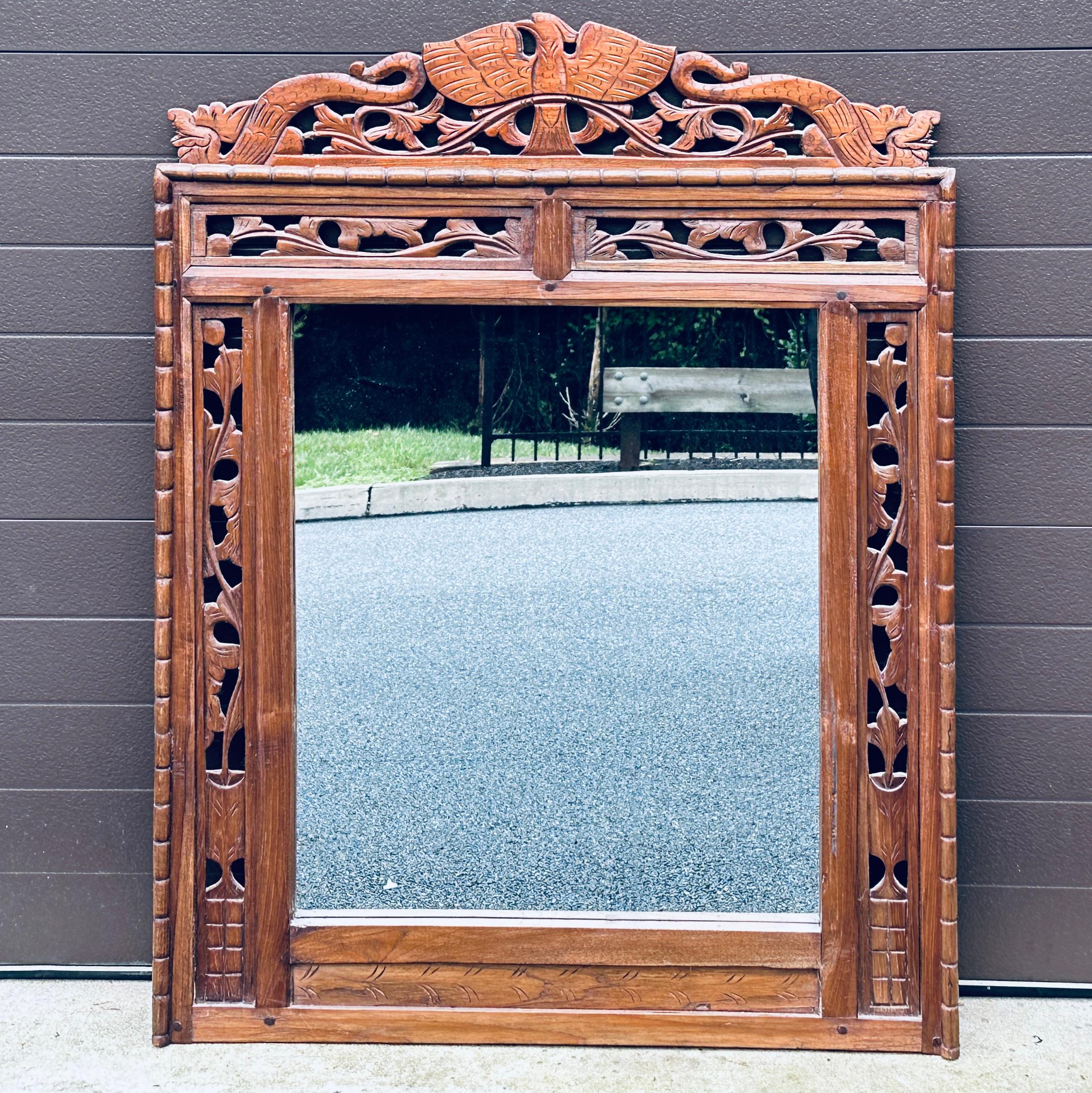 An antique Indonesian hand carved teak wall mirror with openwork and mortise and pegged construction featuring an epic crest of a large bird with serpents on either side. Mirror view 24”w x 27”h. Our client originally purchased this mirror in