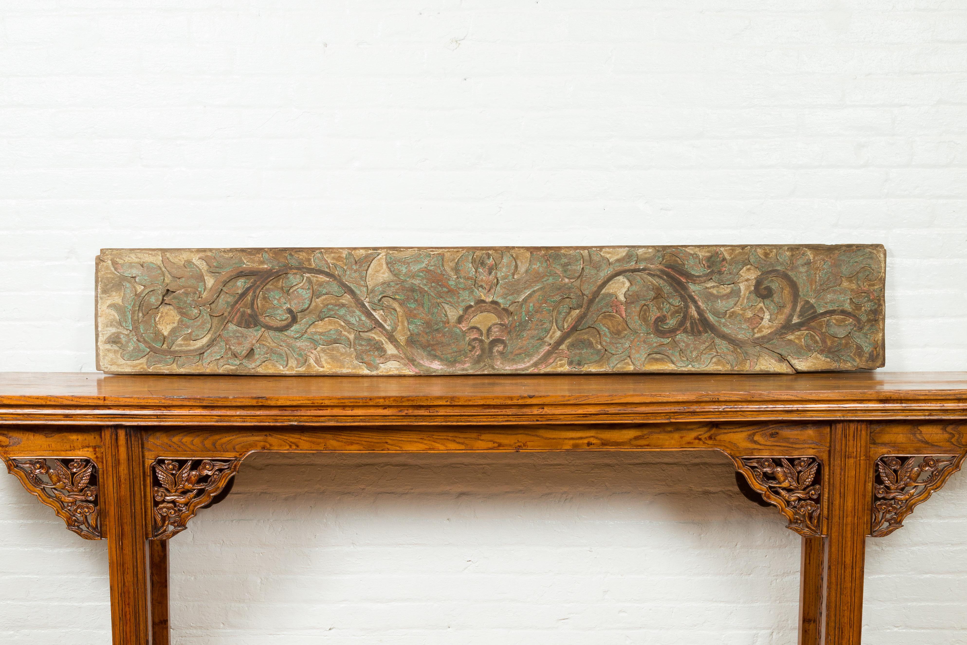 An antique Indonesian painted and carved wooden door panel with scrolling motifs and foliage. Handcrafted in Indonesia, this overdoor panel features a rectangular shape adorned with delicate scrolls supporting a luscious foliage. Carved in low