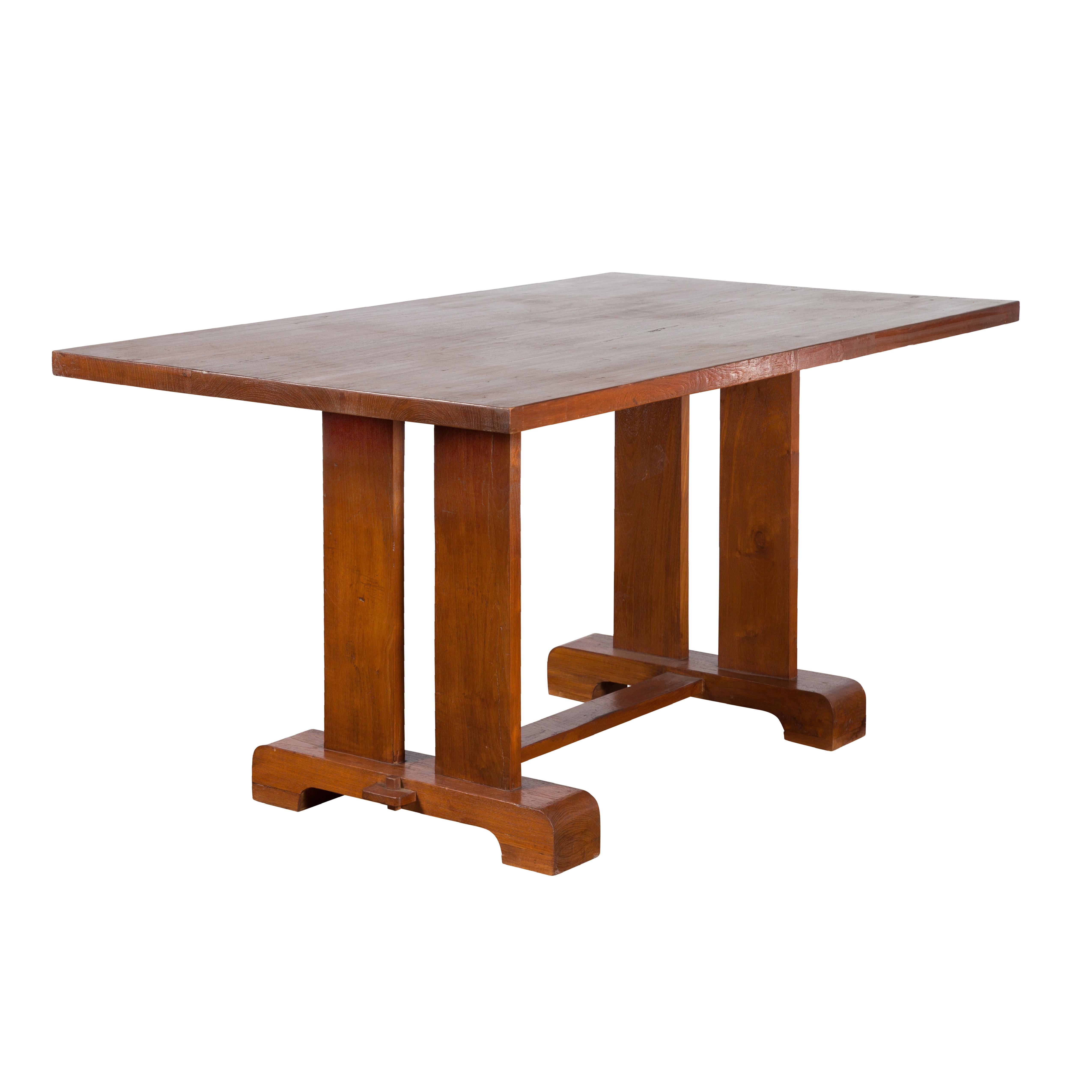 An antique Indonesian teak wood dining table from the early 20th century, with trestle base and brown patina. Created in Indonesia during the early years of the 20th century, this teak dining table features a rectangular top overhanging a simple