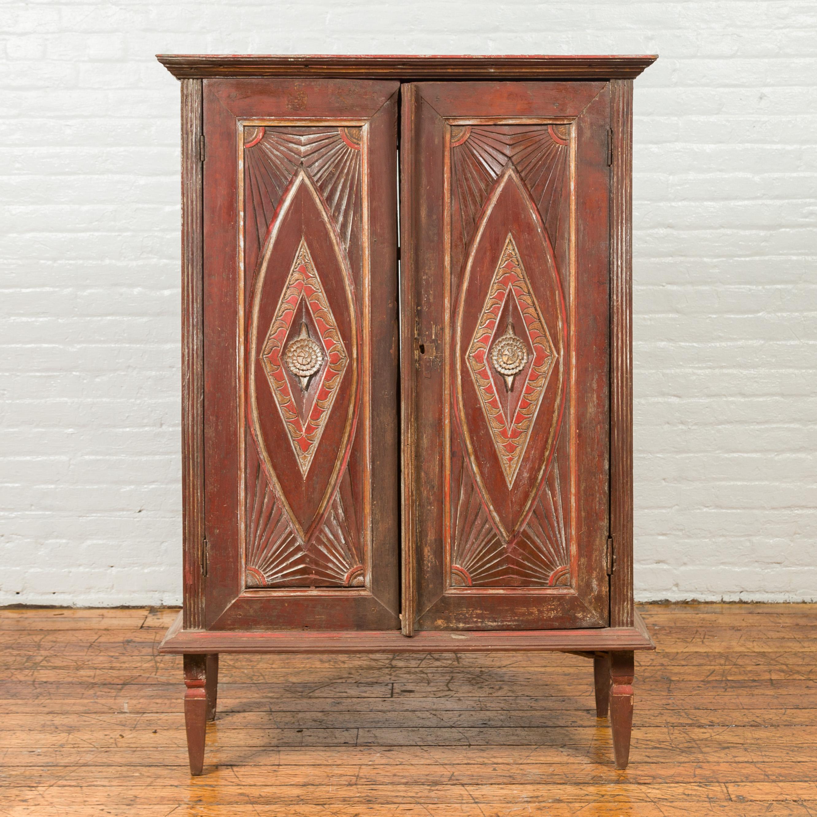 An antique Indonesian wooden cabinet from the 19th century, with carved and painted doors. Born in Indonesia during the 19th century, this wooden cabinet features a beveled cornice sitting above two doors, carved with geometric motifs, sunrays and