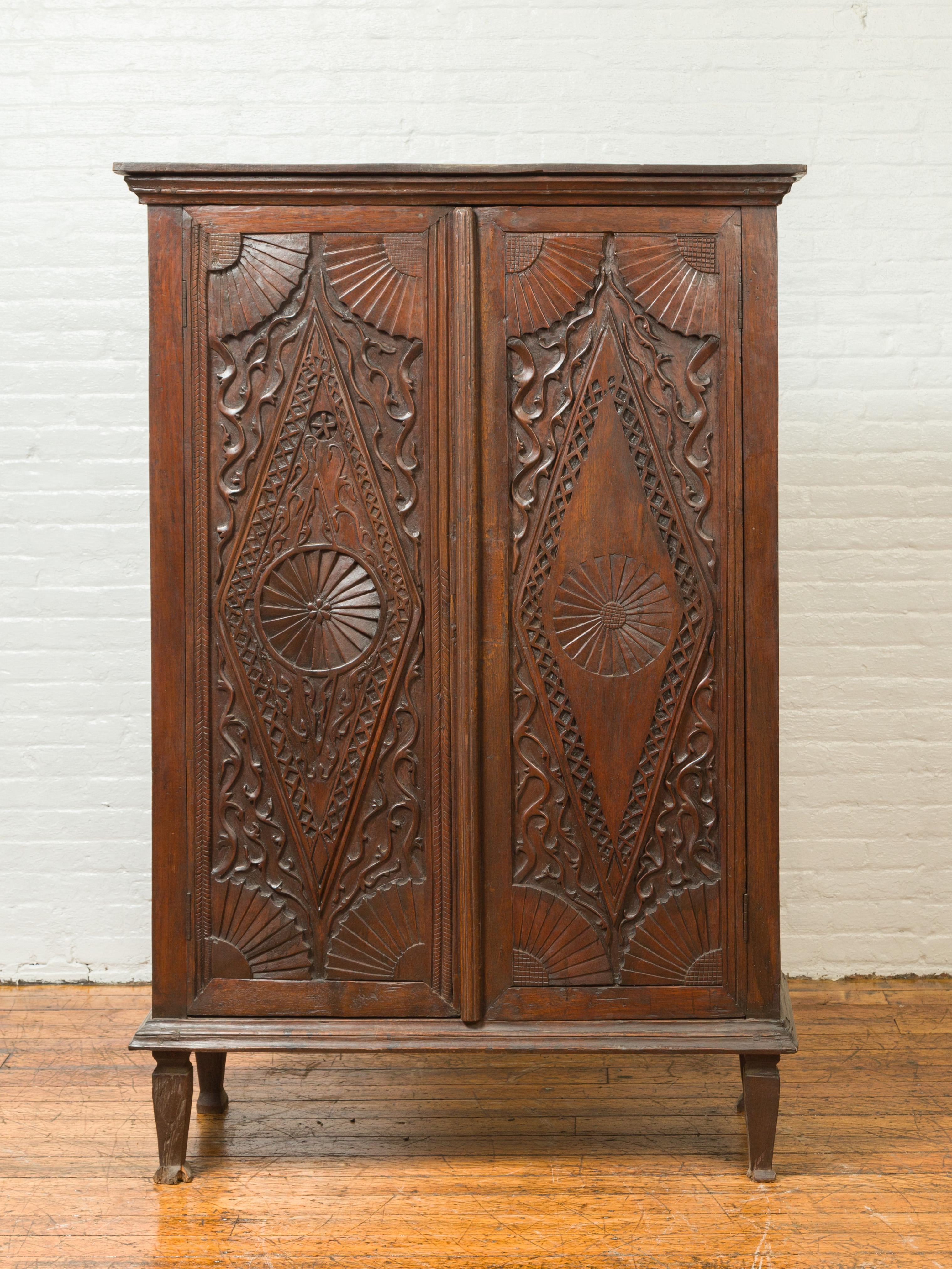 An antique Indonesian wooden cabinet from the 19th century, with carved diamond and radiating motifs. Crafted in Indonesia during the 19th century, this wooden cabinet features a beveled cornice sitting above two intricately carved doors, each