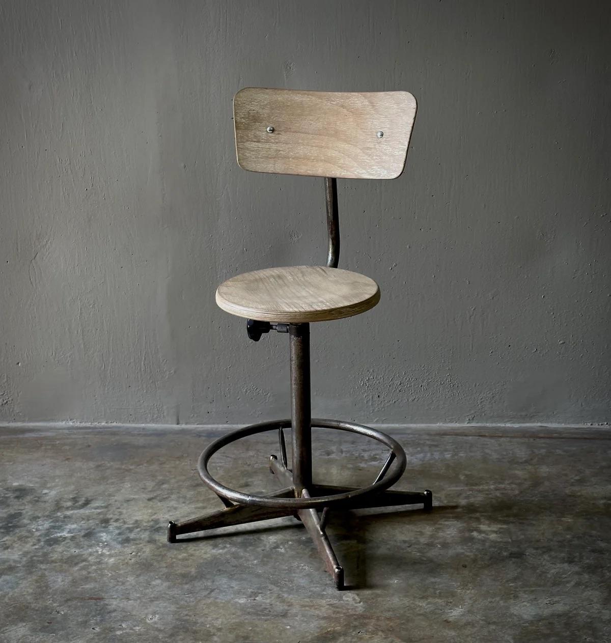 This French industrial swivel stool marries form with function, featuring a minimalist light blonde wood construction paired with an adjustable metal base. The circular seat and curved trapezoidal back are supported by a sturdy pedestal with a ring
