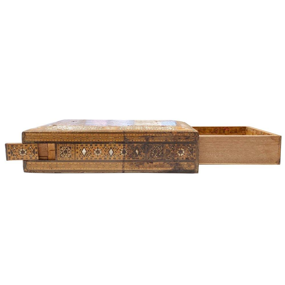 Esempio accattivante di artigianato siriano, questa scatola in legno intarsiato risale al 1960 e presenta intarsi geometrici dettagliati con motivi tradizionali. Al di là del suo fascino decorativo, la scatola include una botola nascosta e un