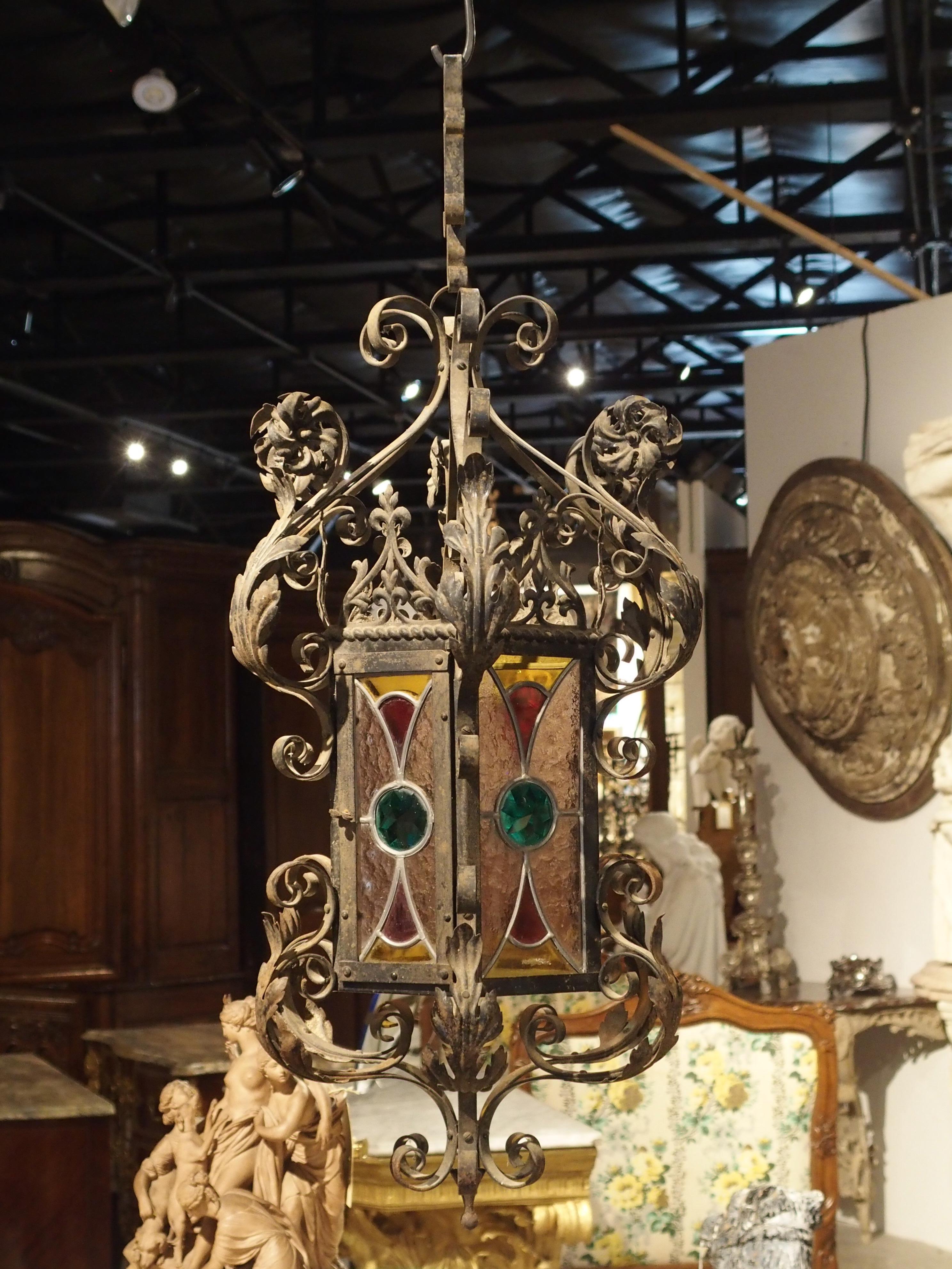 This charming antique French iron and stained-glass lantern from Dijon dates to the early 1900s. The body where the stained glass is has an indented octagonal shape with acanthus leaf C and S scrolls above and beneath it. At the top centre of each