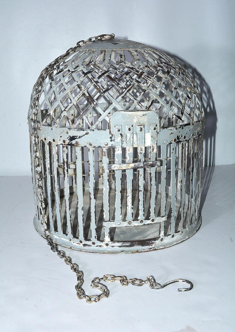 The antique painted iron bird cage has slats and a lattice-work dome. The door slides up and down. There are a wide swing, and a perch to which two iron feeding bowls are attached. The bottom is painted iron as well. The effect is that of a garden