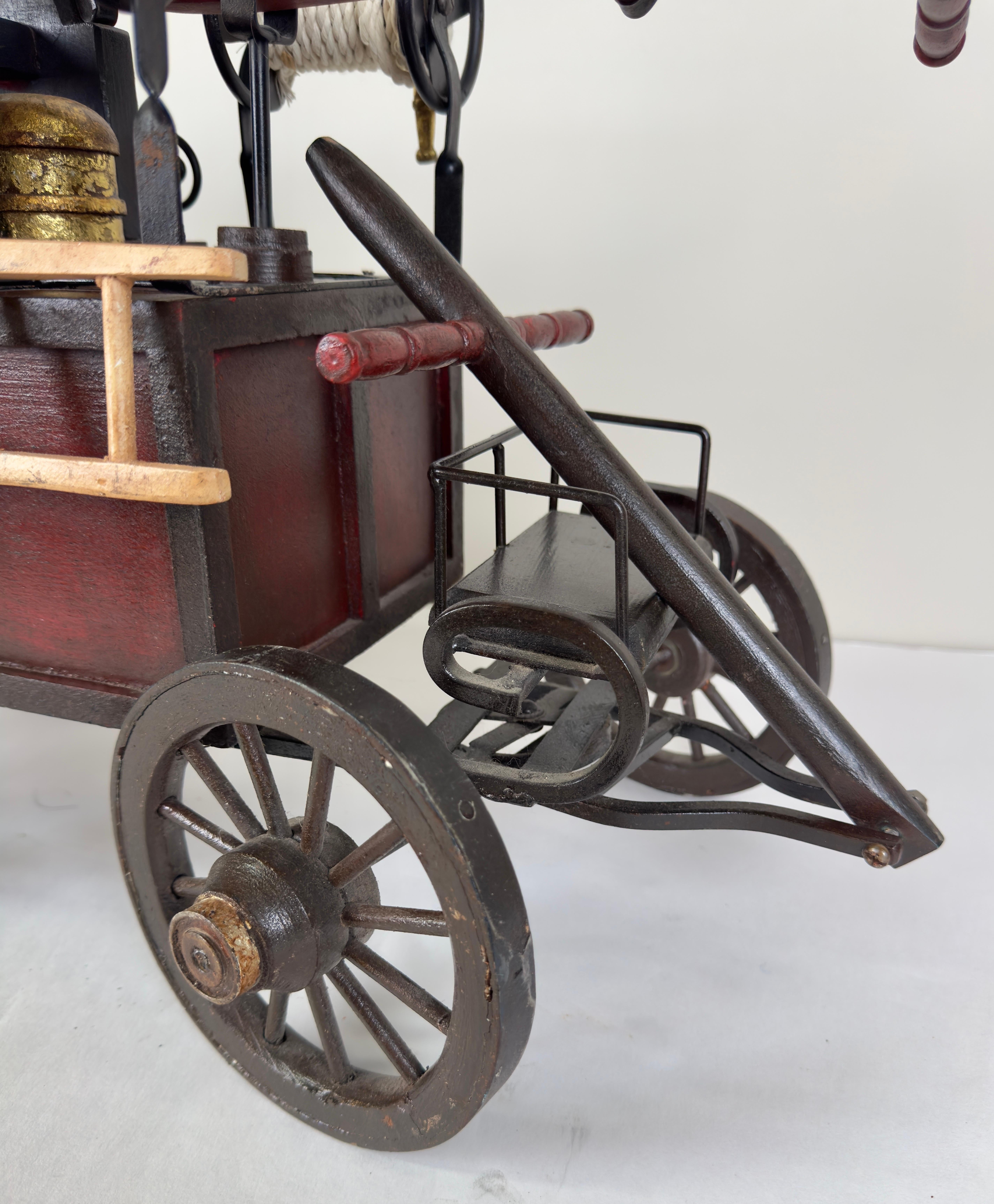 An antique fire engine model or sculpture, crafted from iron and wood, representing a fire engine from the 18th or 19th century. 
The model is mounted on a four-wheeled wooden cart, typical of early fire engines. The wheels are spoked, with iron