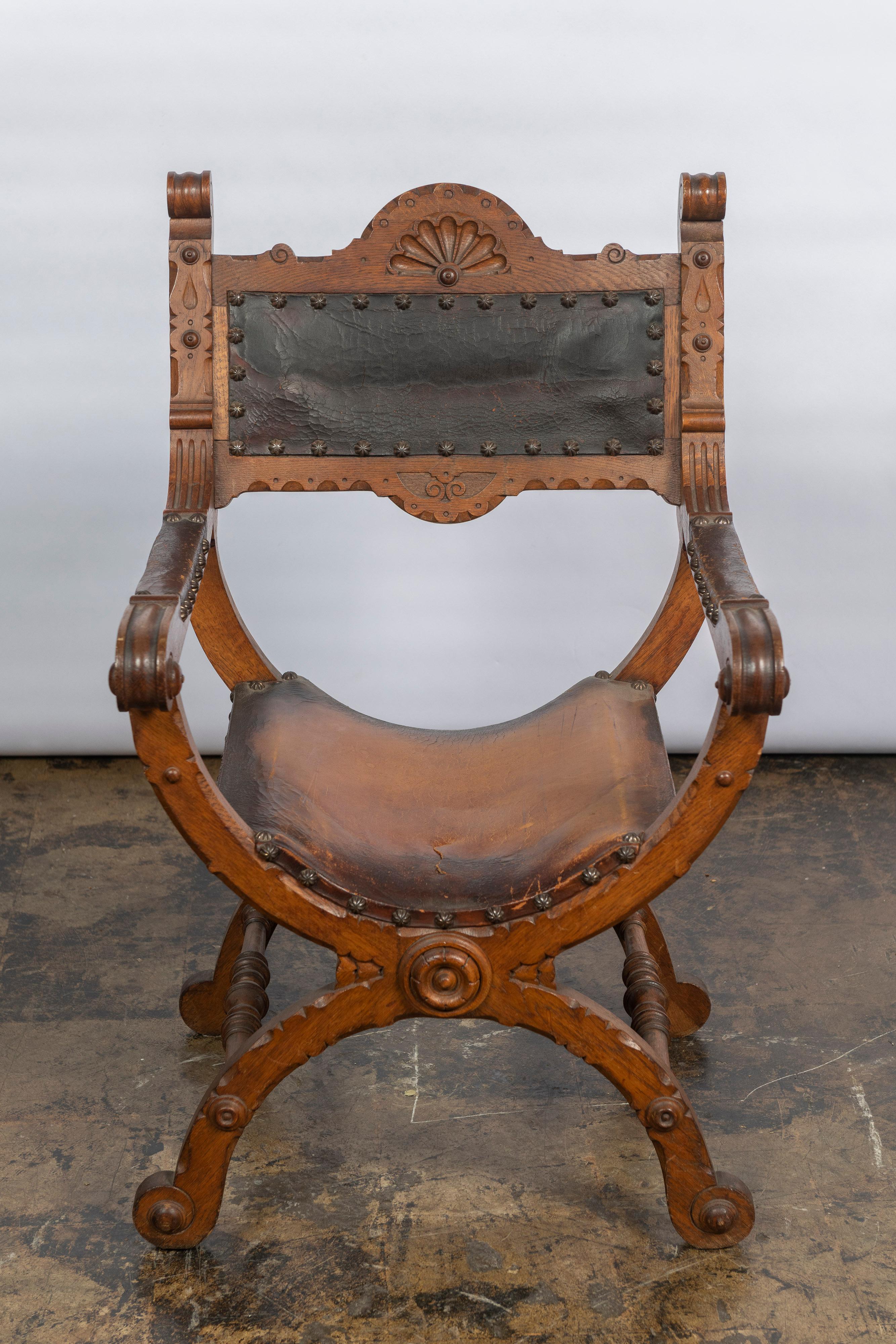 This style of chair is often referred to as a Dante or Savonarola chair, made of carved wood and heavy saddle leather. The frame is X- shaped and the chair is decorated with detailed carving across the rest arms, back and in the frontal center of