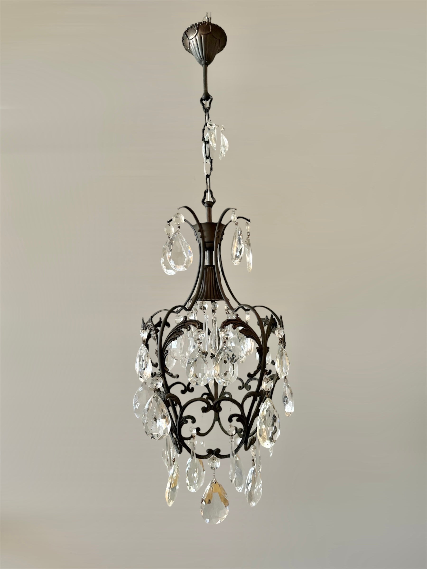 Un élégant lustre italien des années 1950, fabriqué en laiton bronzé foncé et orné de gouttes de cristal de Bohême de haute qualité. Le design en cage ouverte met en valeur les volutes ornées, tandis que les cristaux facettés capturent et reflètent