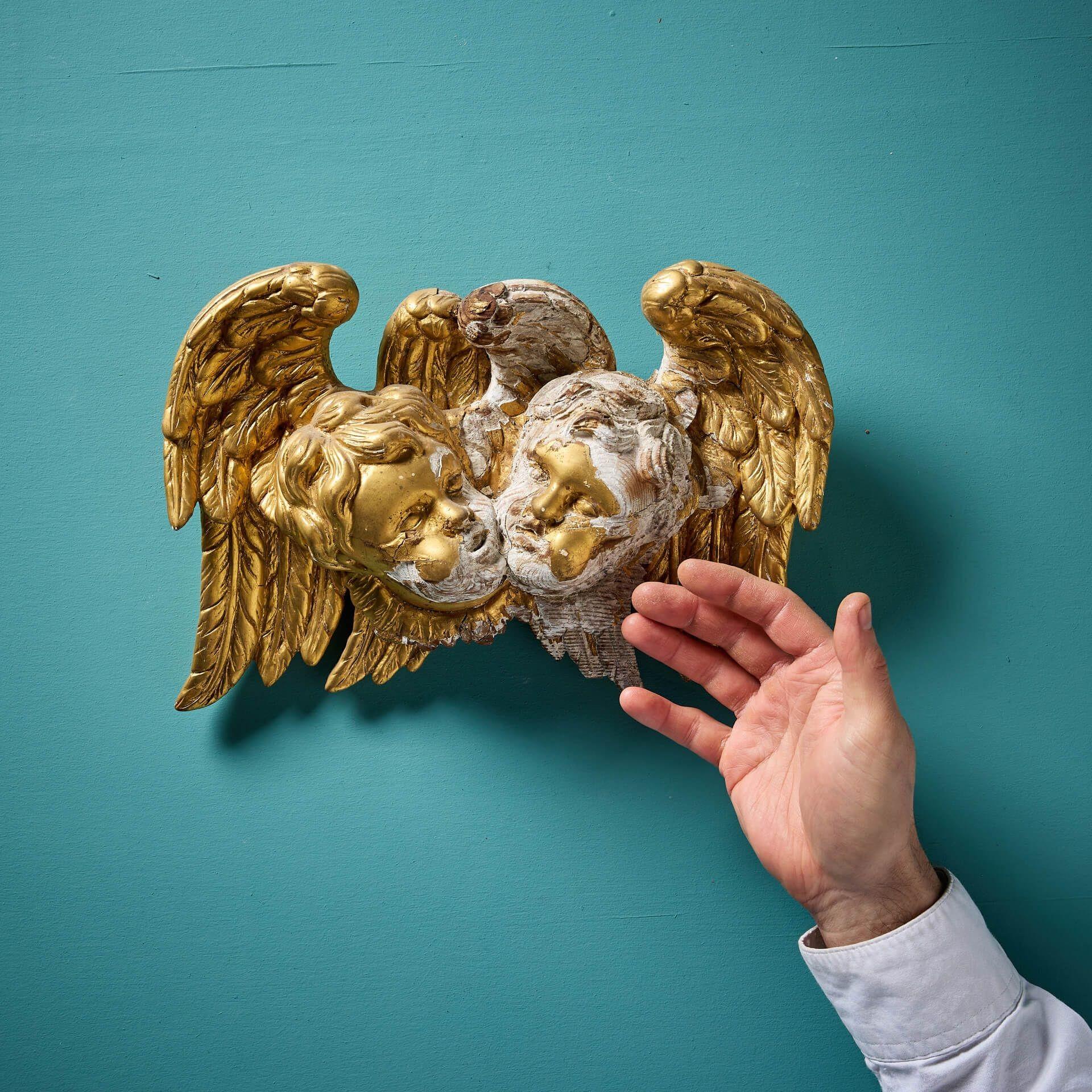 A charming carved and gilded antique putti pair, originating from late 18th / early 19th century Italy, perfect for hanging on an interior wall. Carved in pine and gesso, the piece depicts two cherub masks, each before a magnificent pair of angelic