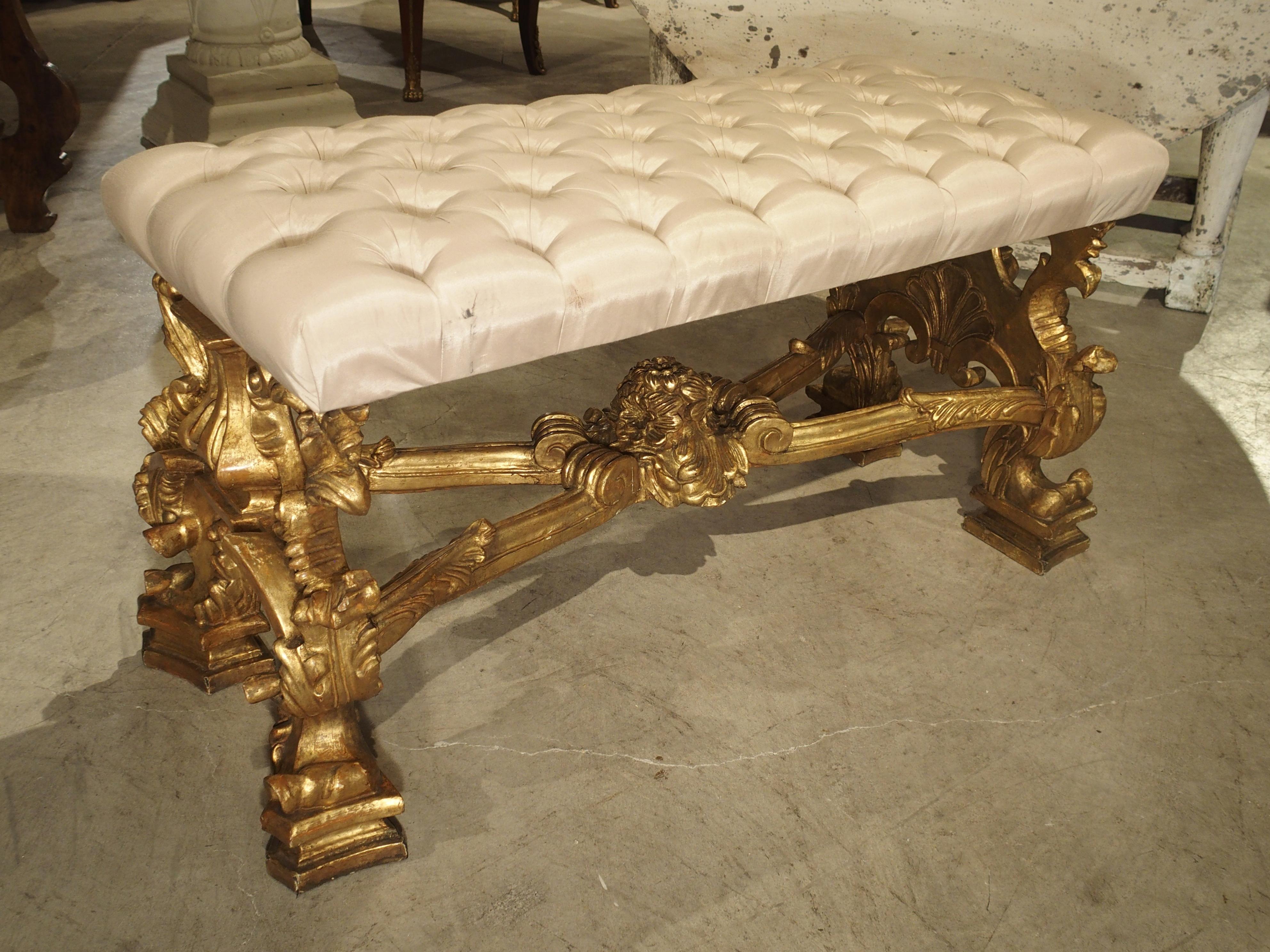 This luxurious giltwood Italian bench is in the Rococo style with an attached tufted seat cushion in a cream colored silk. The gilded legs have motifs of scrolling and gadrooned acanthus leaves with stylized shells on the apron at either end. There