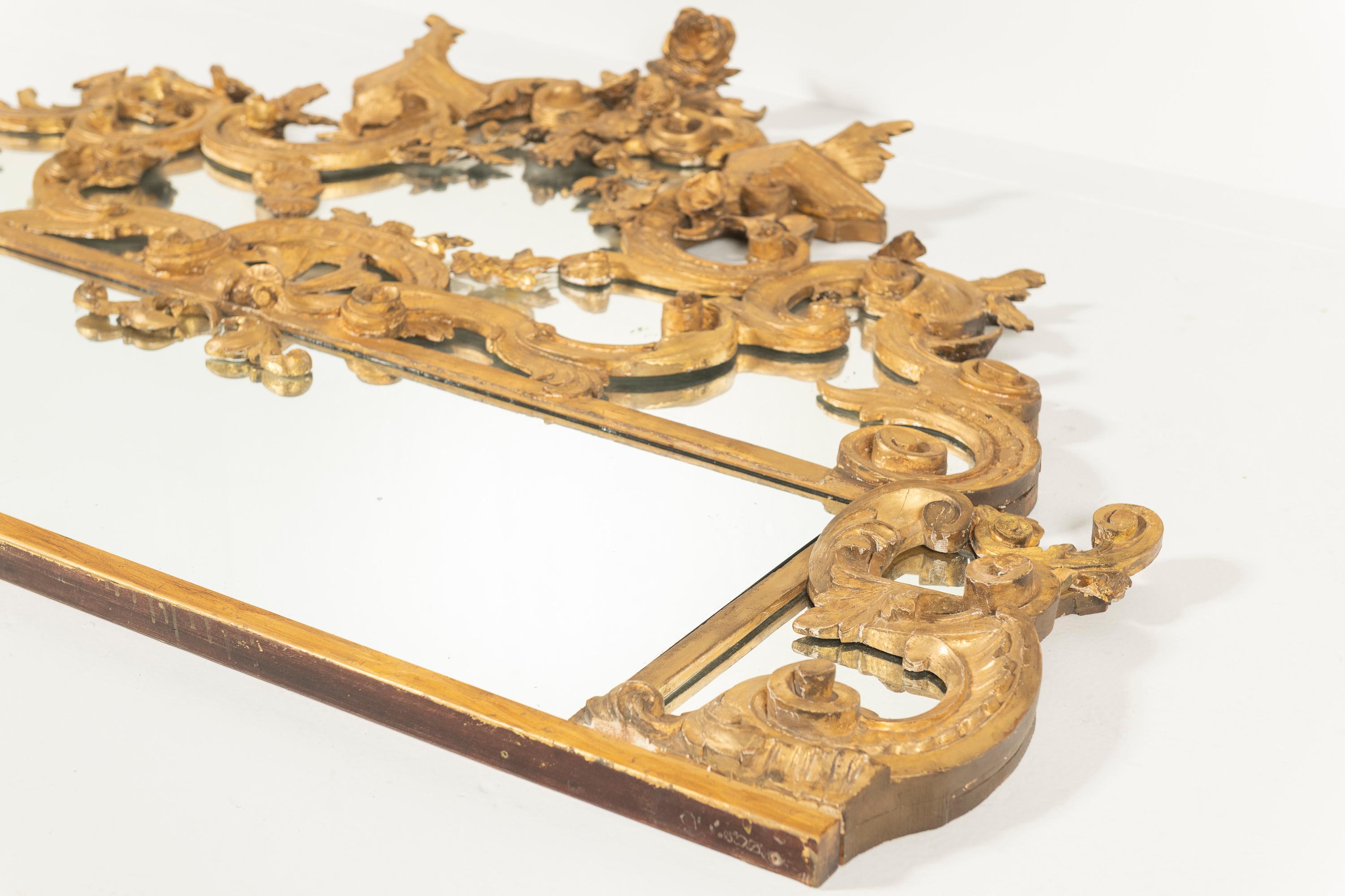 This large Antique Italian Giltwood Mirror has an elegant, decorative design with intricate scrollwork and floral motifs. The gilded frame features a symmetrical arrangement of curved details, giving it a refined, classic look. The mirror is divided