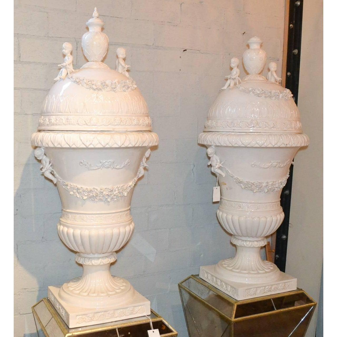 Stunning pair of large Italian glazed porcelain capped urns. Having beautifully intricate floral swag detailing, charming putti adornments, and wonderful glazed patina. Beyond chic for numerous designs!