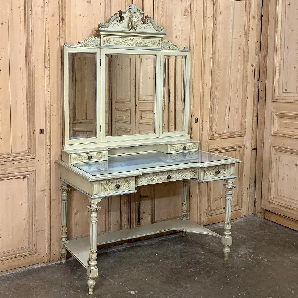 Antique Italian Louis XVI painted vanity features a strong carved crown overlooking the tripanel folding mirror below, which in turn rests atop a pair of drawers and cubby hole, and again which rests upon the main glass topped surface. Carved floral