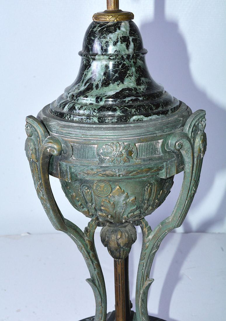 The neoclassical table lamp is in the shape of an ancient Roman lighting fixture. A grey & green-veined dark marble base and upper body is set into a bronze circle set above tripod splayed leaf motif legs. The lamp base is made of the same marble. 