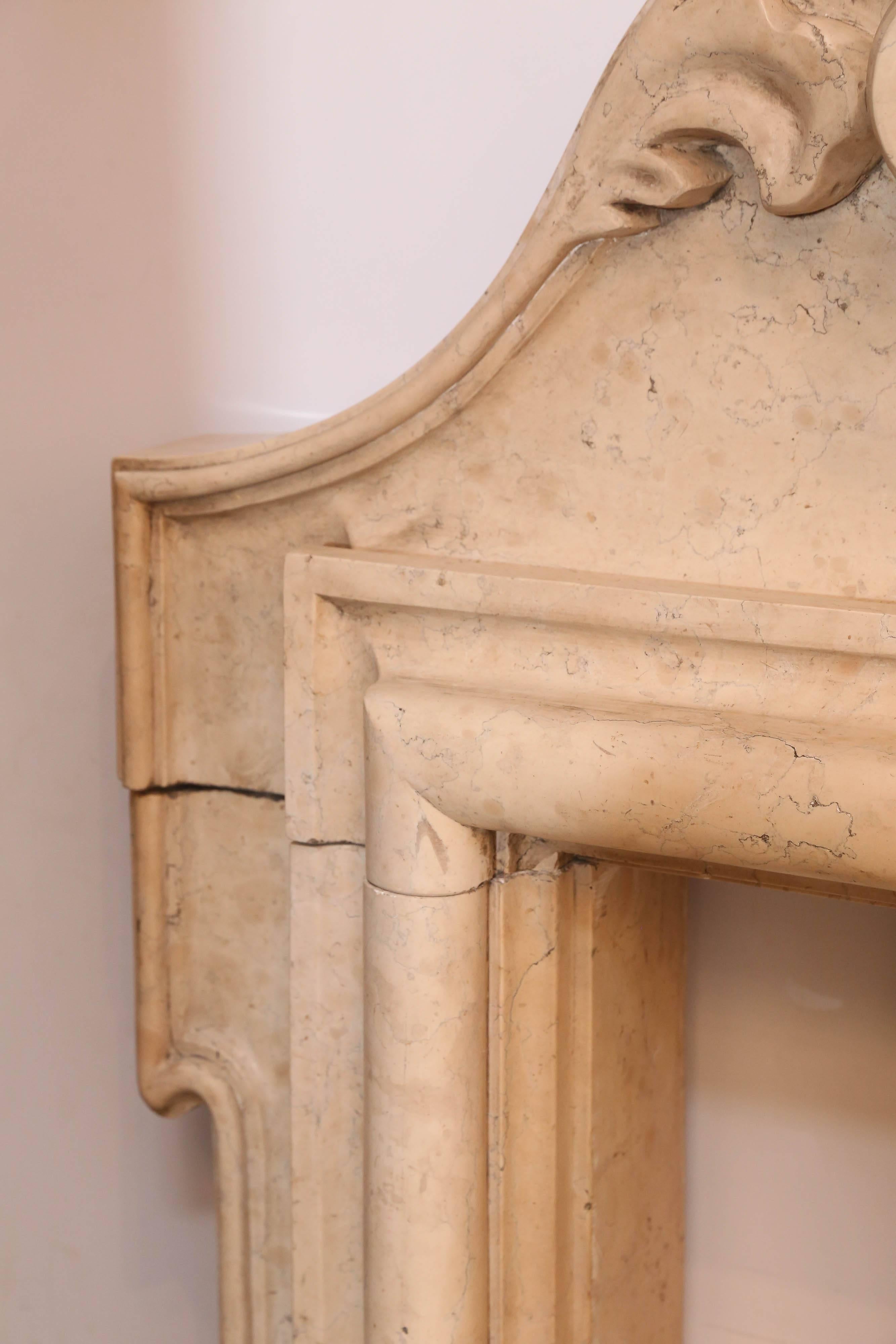 Antique Italian fireplace is carved in a neutral marble with no veins.
Large cartouche is on top with two decorative carvings on either side.
Mantel sides have a simple carving on top of
with a more decorative carving on the bottom corners.
 